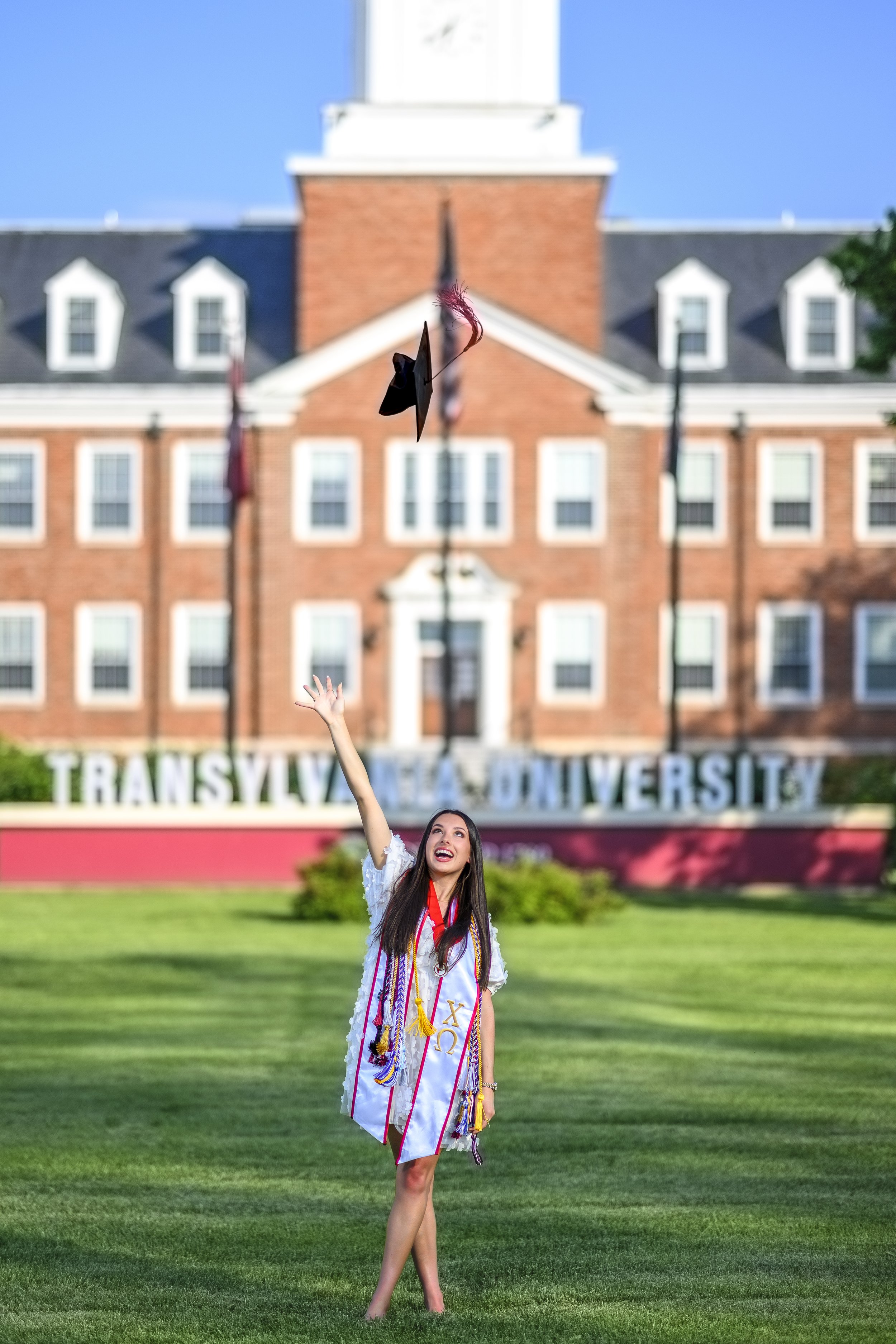 A young woman with long dark hair, wearing a graduation gown and stole with honor cords, is standing on a grassy area in front of Transylvania University. She is tossing her cap into the air, smiling, with her hand raised. The background features a b
