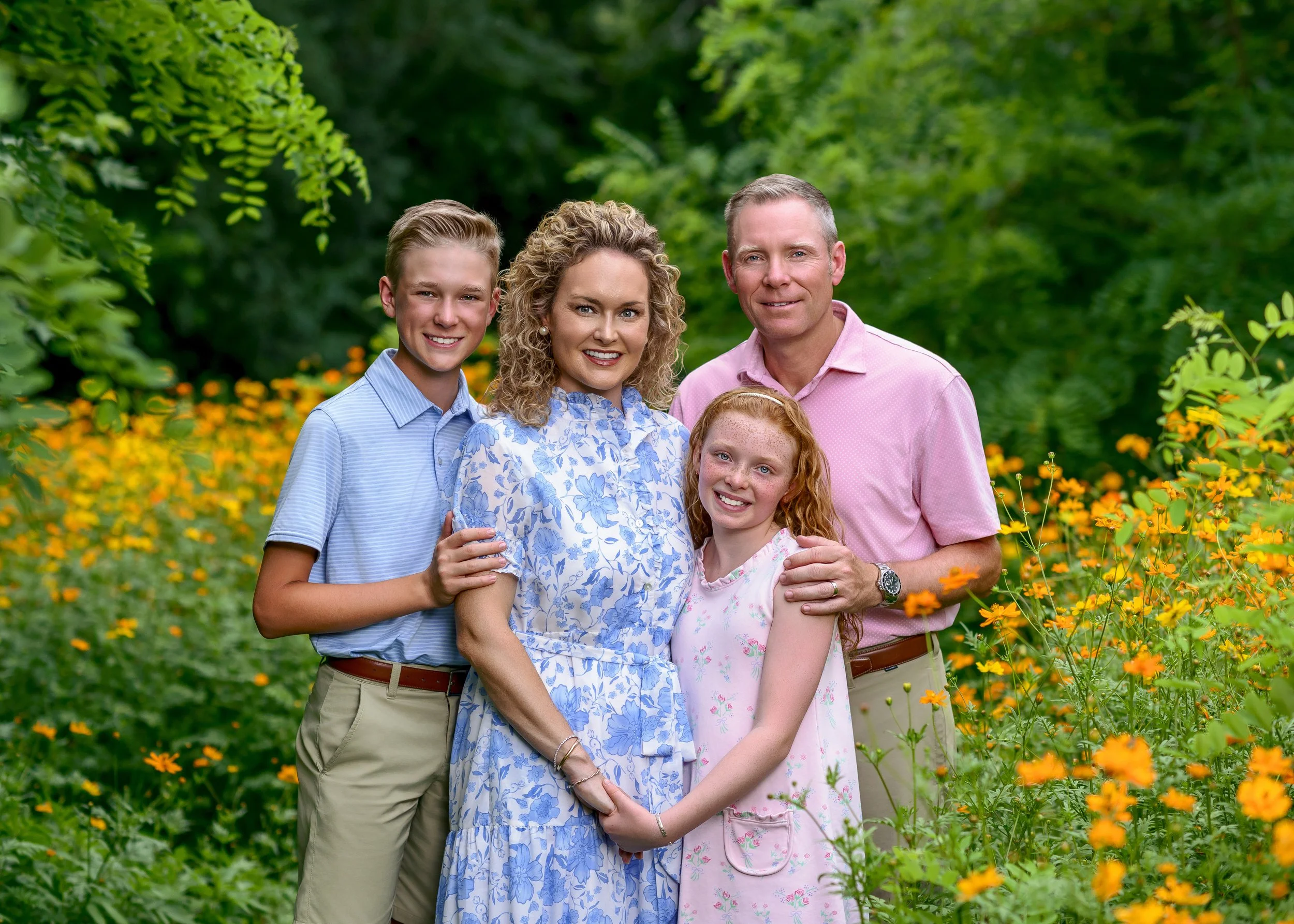 A family of five standing together outdoors among yellow flowers and green foliage, smiling at the camera.