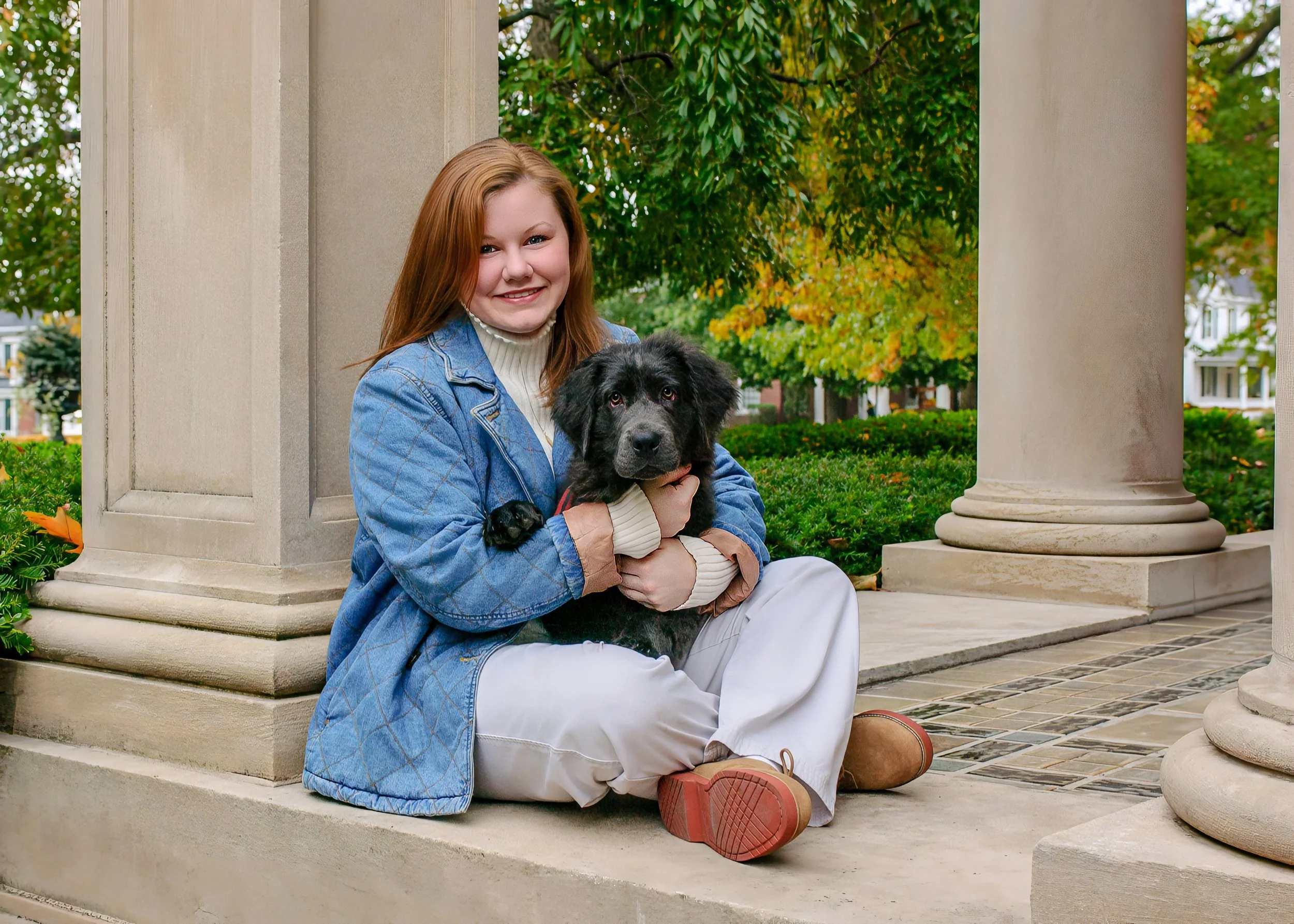 A young woman with red hair wearing a denim jacket and white pants sitting on the steps of a stone building, holding a black puppy with white markings. The background shows green and yellow trees and a well-maintained yard.