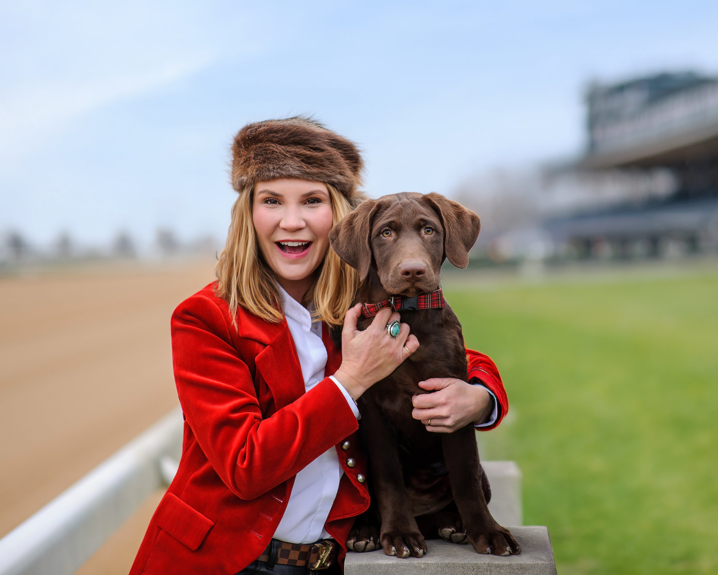 A woman in a red blazer and white shirt is smiling and holding a young chocolate Labrador Retriever with a red and black plaid collar on a bench outdoors.