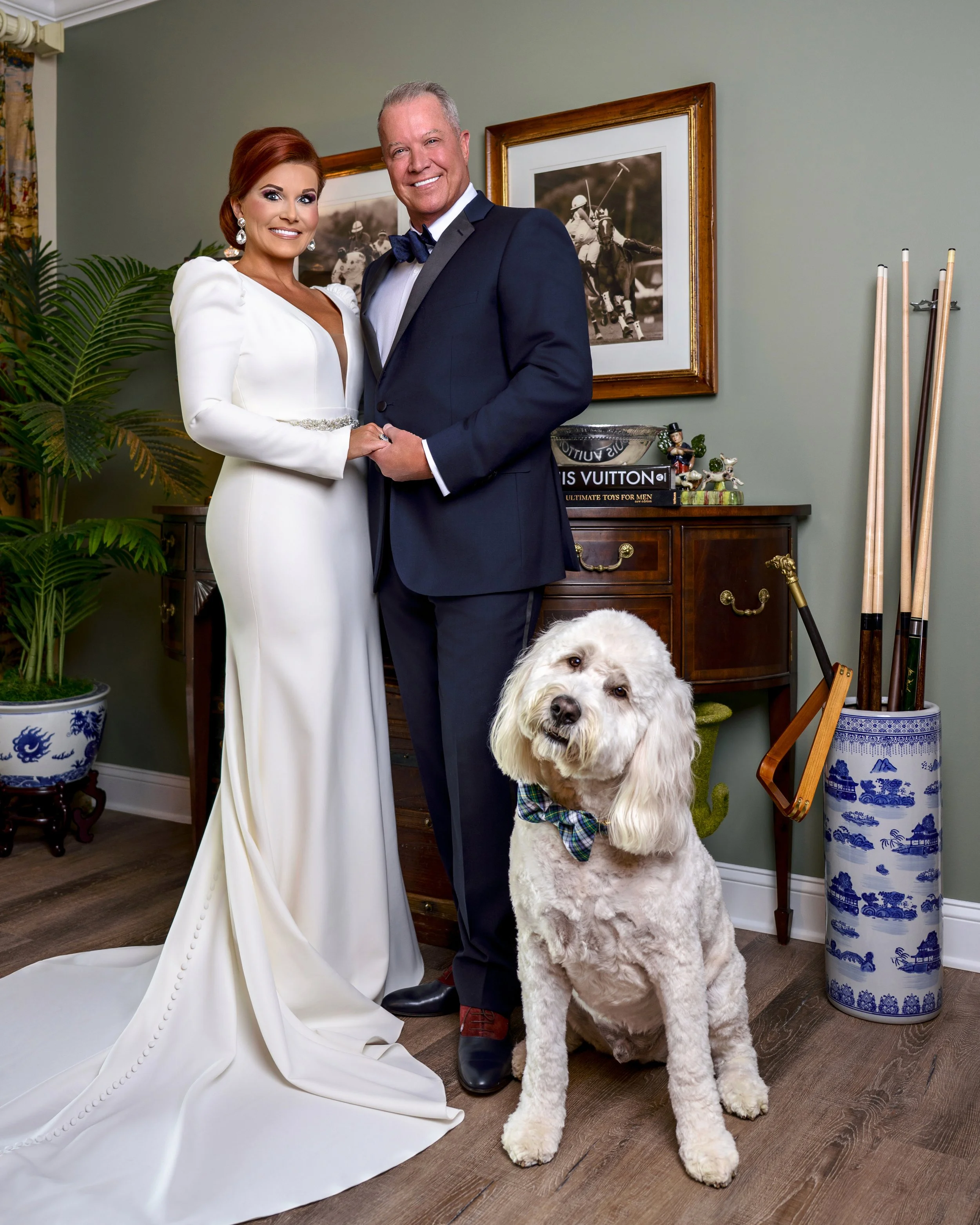 A couple dressed in formal wedding attire, with the woman in a white gown and the man in a dark suit and bowtie, standing indoors with a large dog wearing a plaid bow tie in front of them. Behind them are framed photographs of polo players, a wooden 