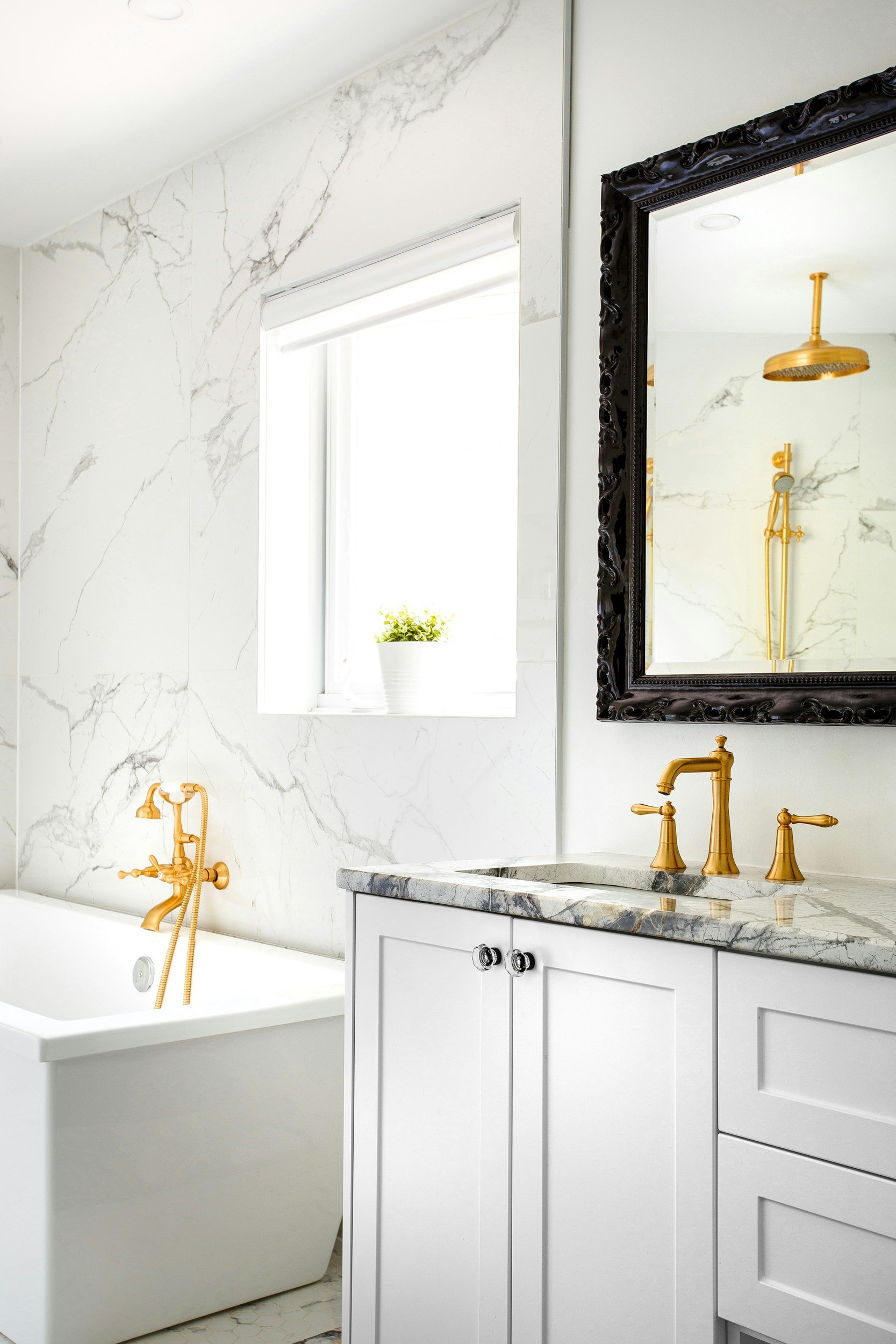 A luxurious white bathtub with gold fixtures next to a marble vanity with gold fixtures, a large mirror with a black ornate frame, a window with a small potted plant, and a shower with a gold shower head and handheld spray.
