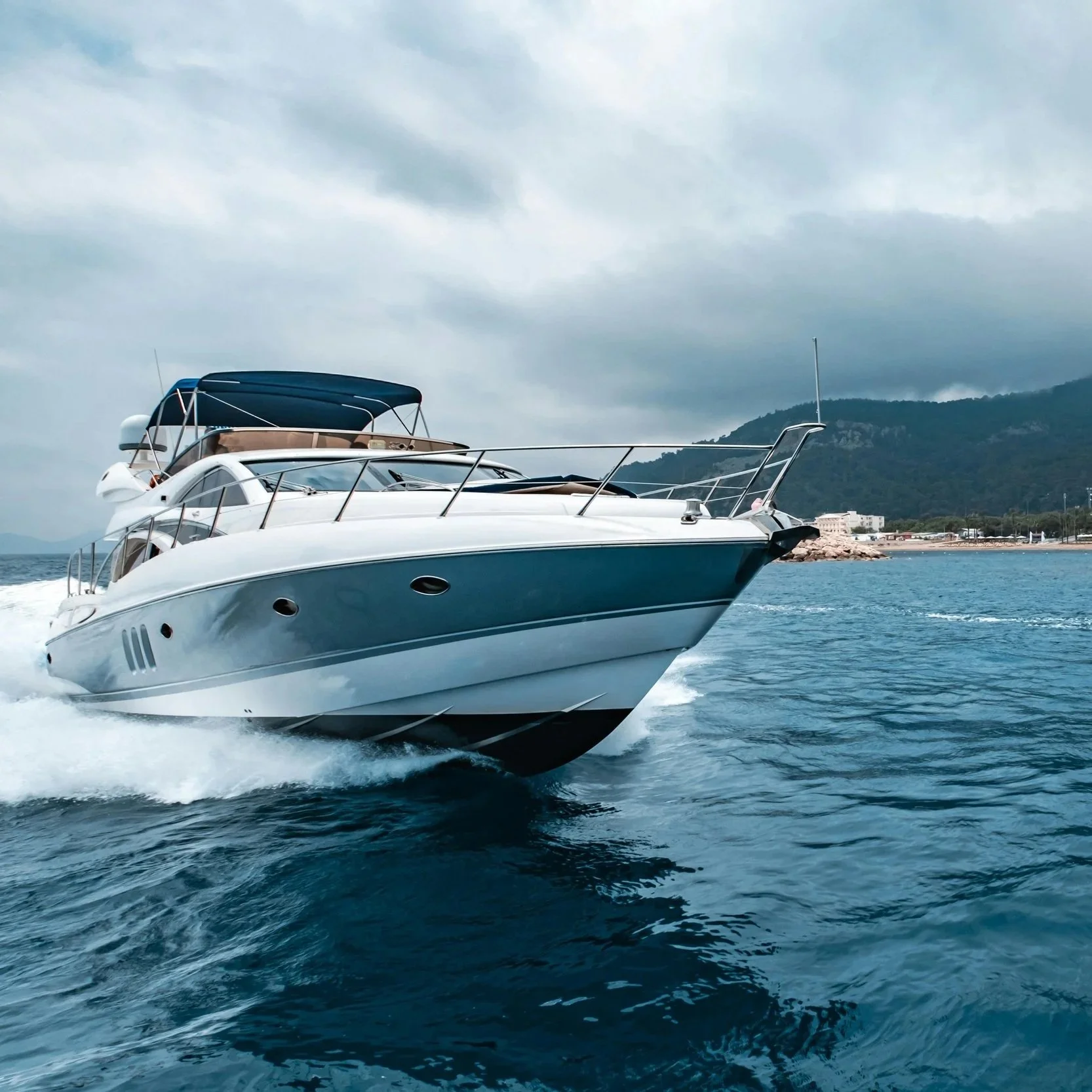 A white yacht moving quickly through the water near a shoreline with buildings and a mountain in the background, under a cloudy sky.