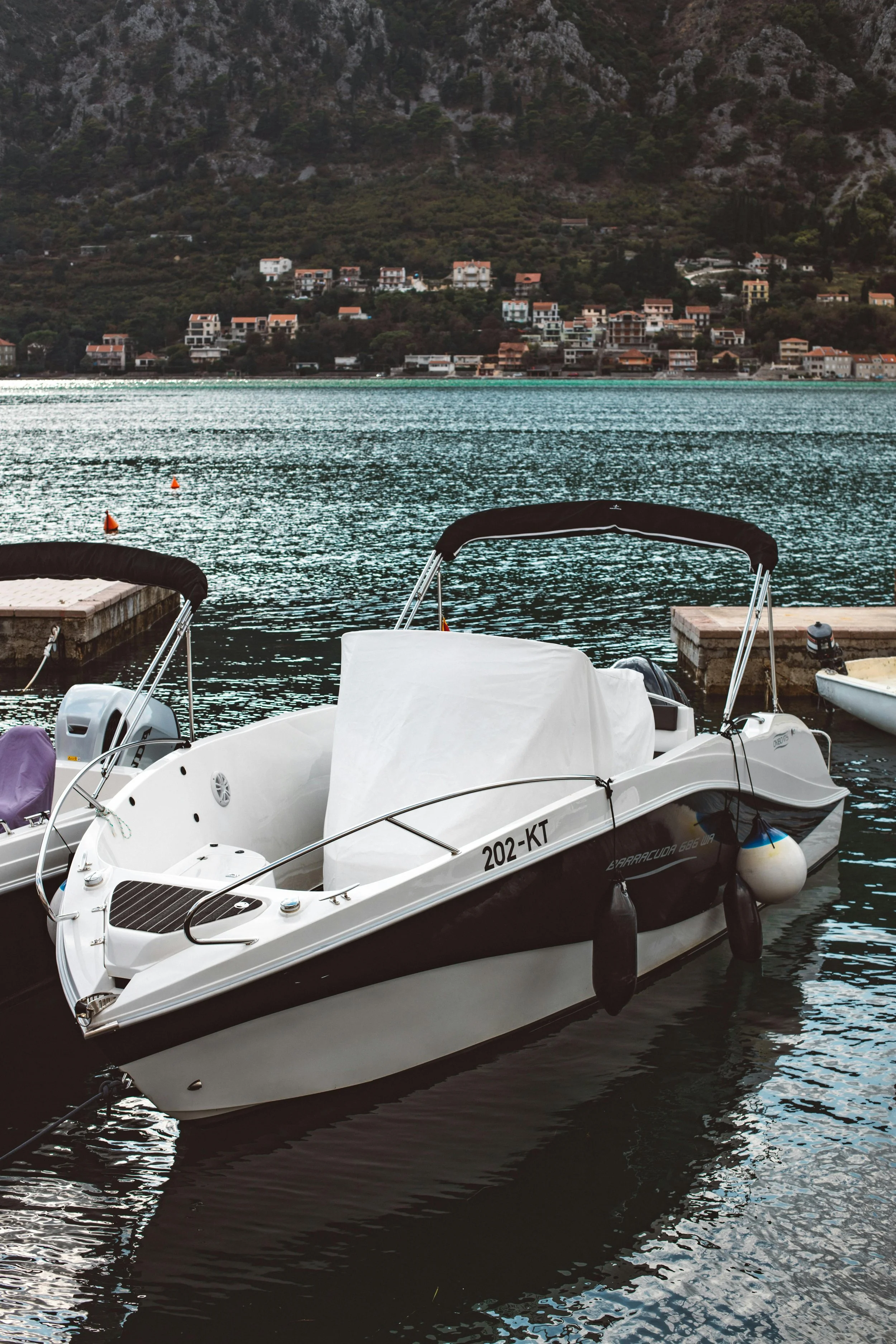 A white motorboat docked at a marina on a lake with mountain and residential buildings in the background.