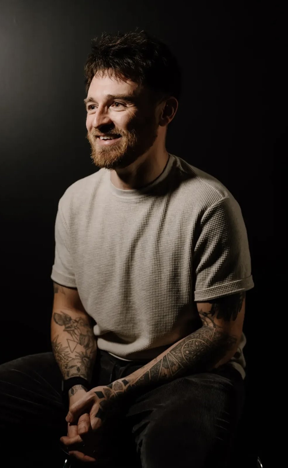 A smiling man with a beard and tattoos sitting against a dark background, wearing a light-colored t-shirt.