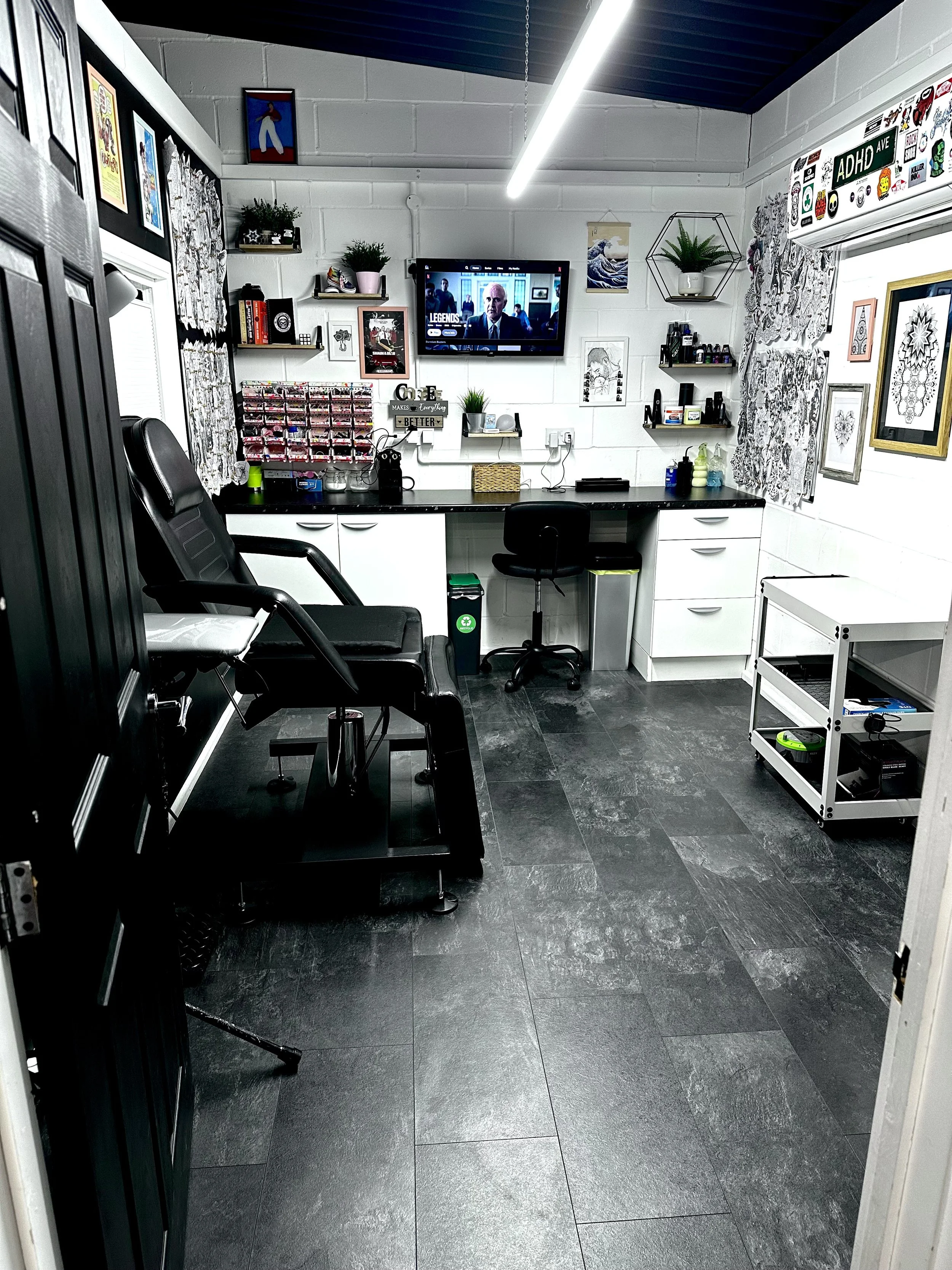 A small, organized art studio or craft room with a black and white color scheme, featuring multiple shelves with art supplies, framed artwork on white brick walls, a wall-mounted television, and a black desk with a swivel chair. The room has dark gray tiled floors, window curtains with black and white patterns, and various decorative plants.