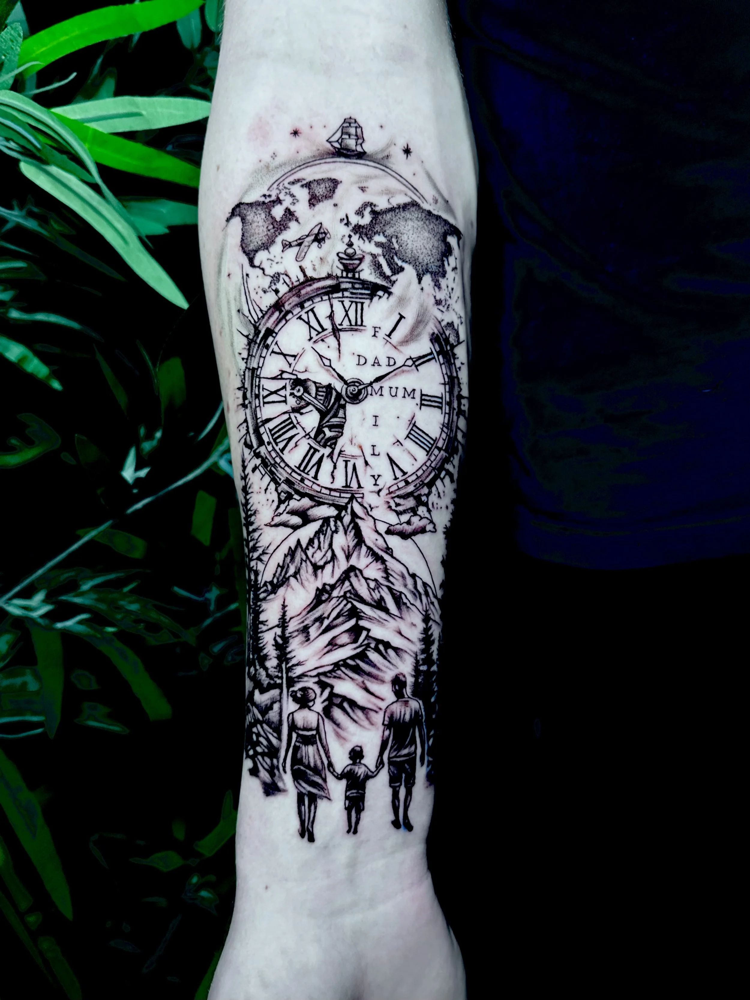 Tattoo on forearm depicting a world map with an airplane, a sailing ship, and a clock showing around 10:10, with the words 'Father' and 'Mummy,' mountains, and people holding hands.