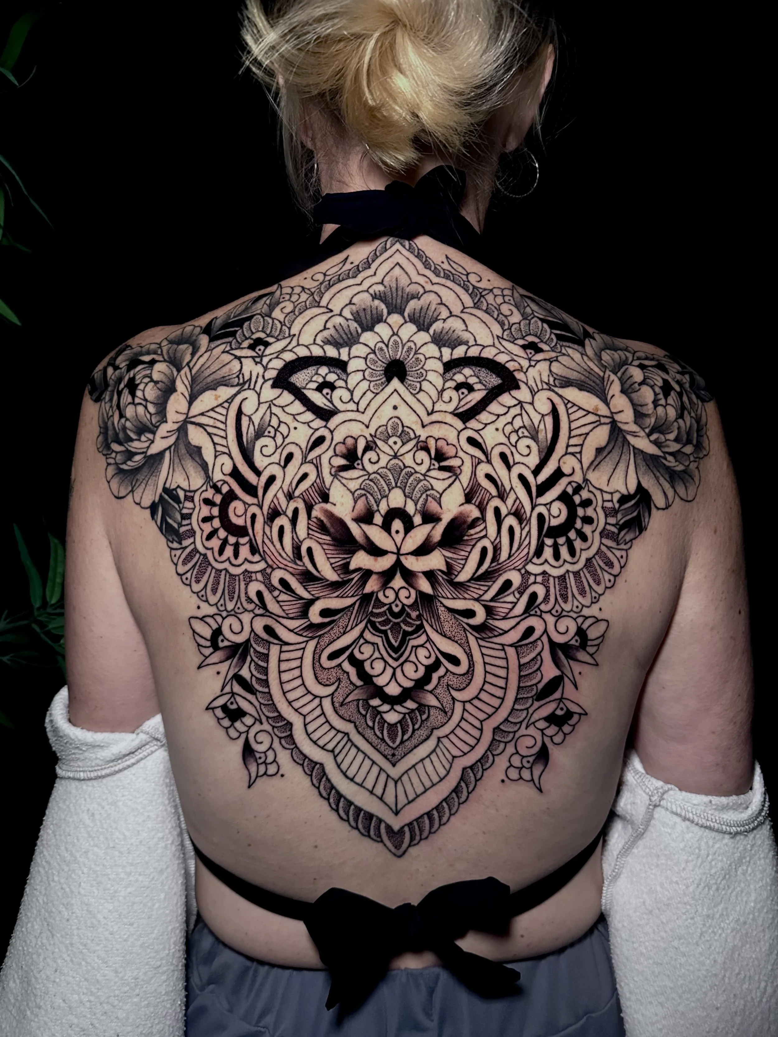 A woman with blonde hair and hoop earrings showing a large, intricate black and grey tattoo on her upper back and shoulders, with part of her white sweater sleeve visible on the left.