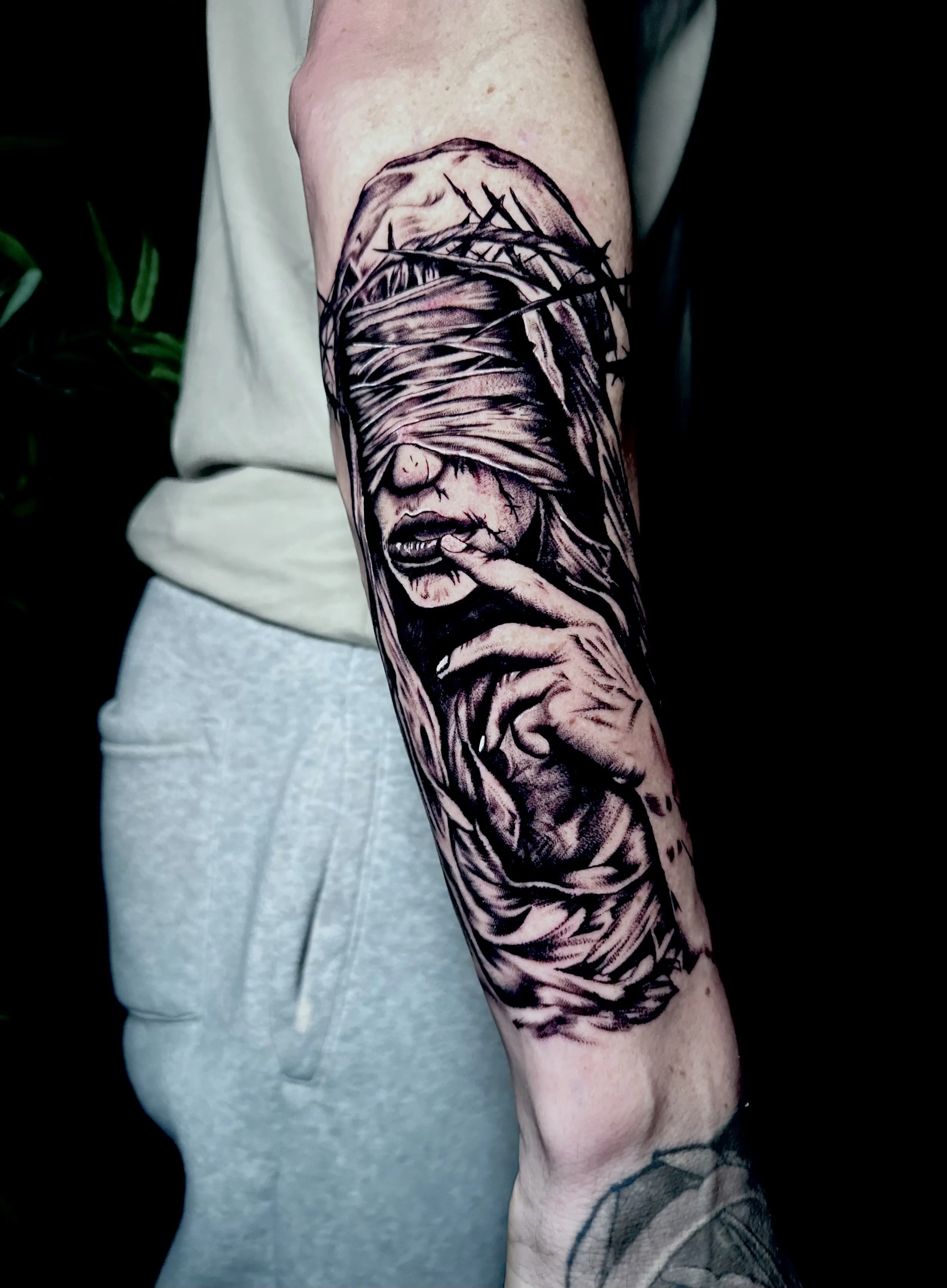 A detailed black and gray tattoo of a woman's face with her eyes blindfolded by bandages, on a person's forearm.