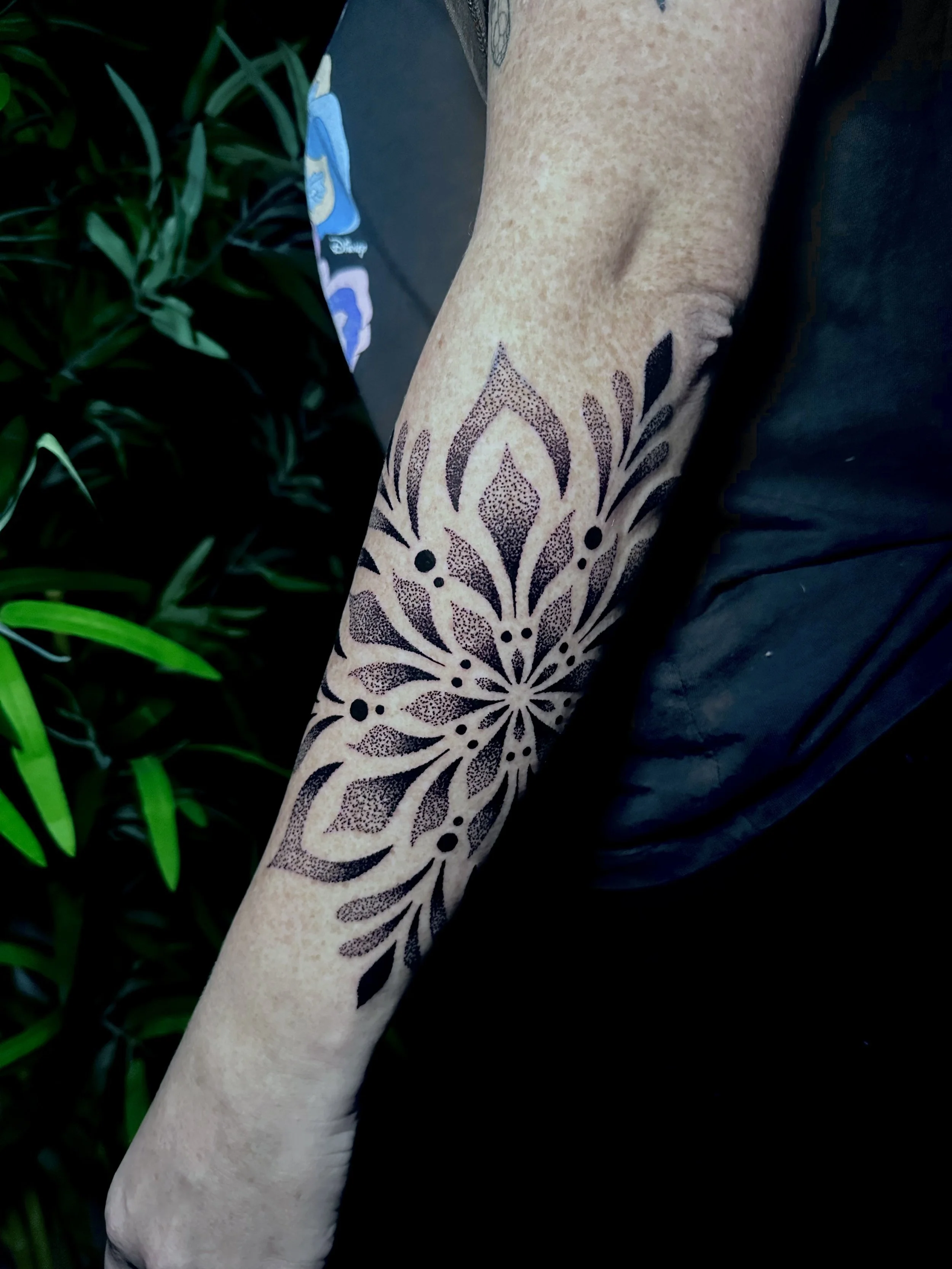 A person's arm with a large black floral tattoo extending from the wrist to the elbow, with green foliage in the background.