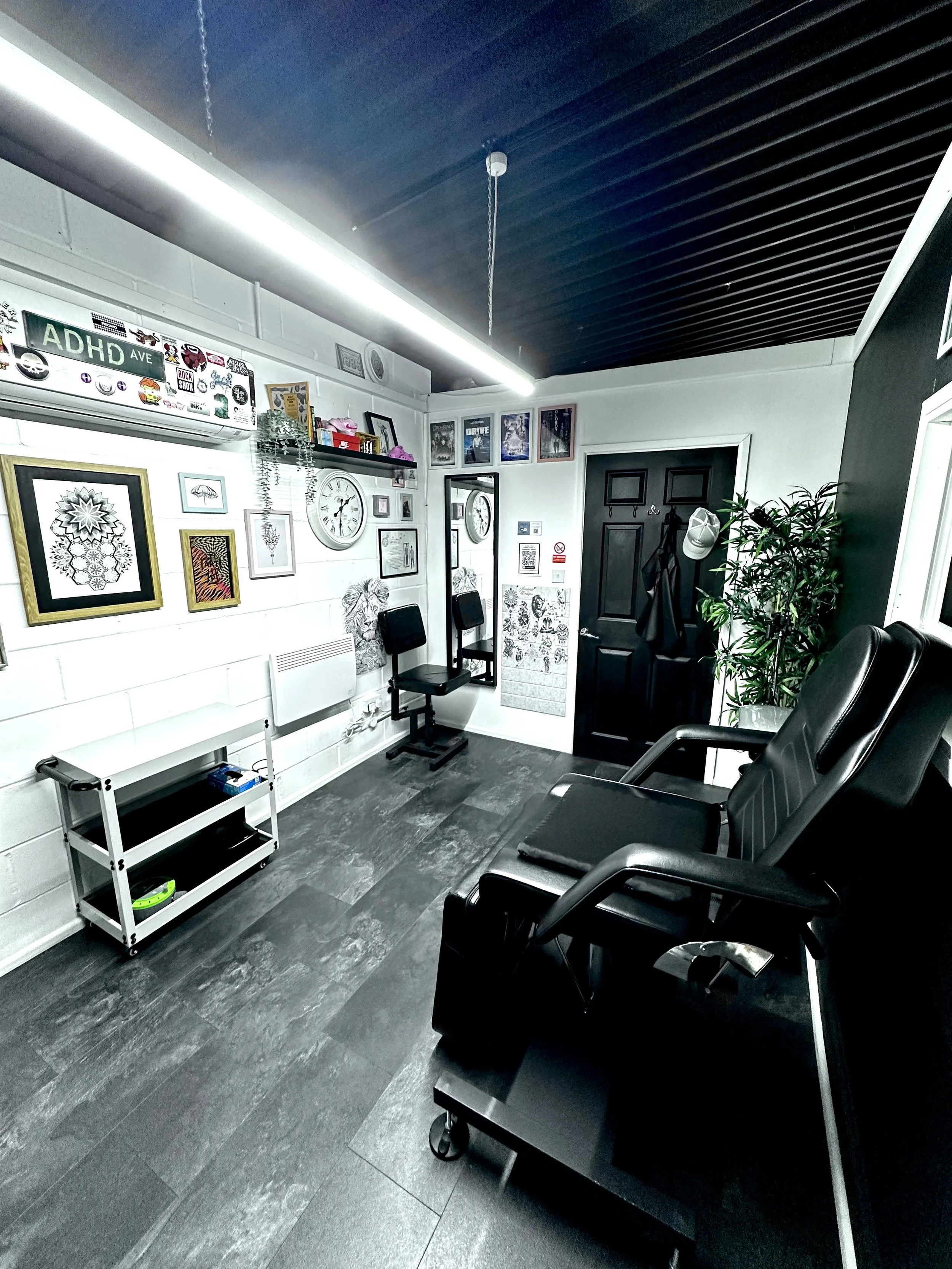 Interior view of a modern waiting room with black chairs, a plants, a white wall decorated with framed pictures and a large clock, and a ceiling with dark wood slats and a strip of LED lighting.