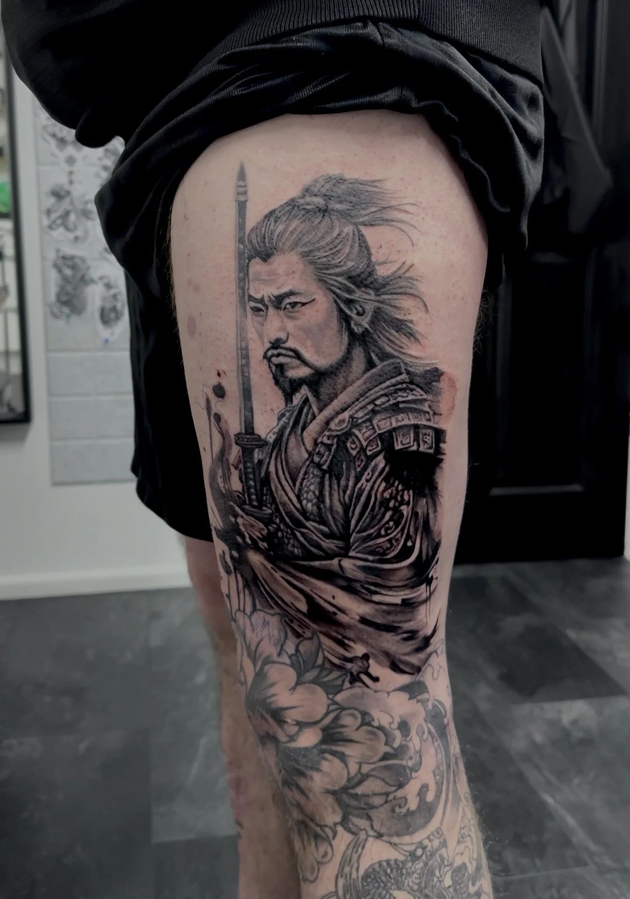 Black and gray tattoo of a warrior with long flowing hair holding a sword on a person's thigh, with a background of clouds.