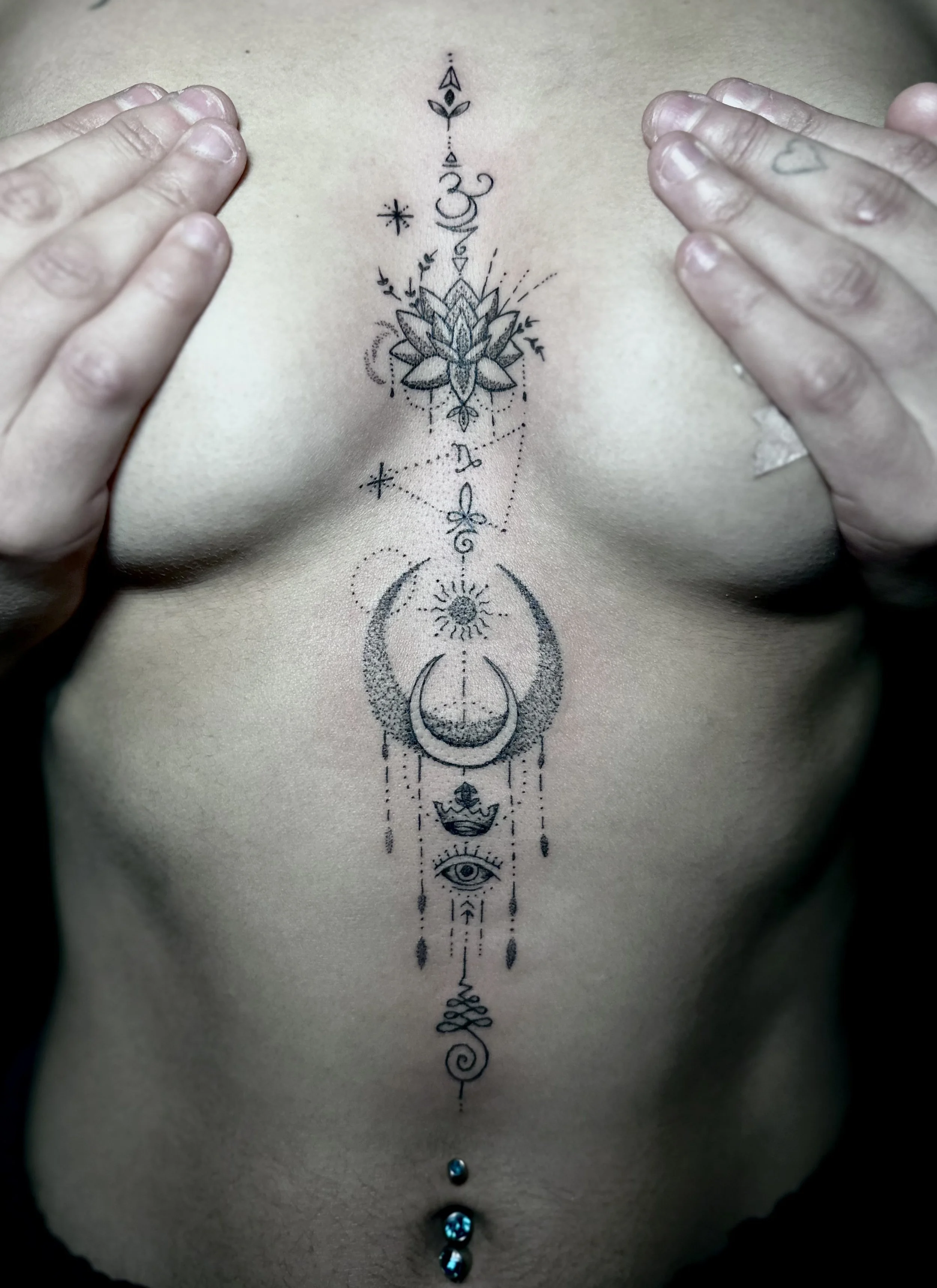 A person's torso with a vertical tattoo featuring various symbols, including a lotus flower, moons, a sun, and an eye, with hands on either side of the tattoo, and three belly button piercings below the tattoo.