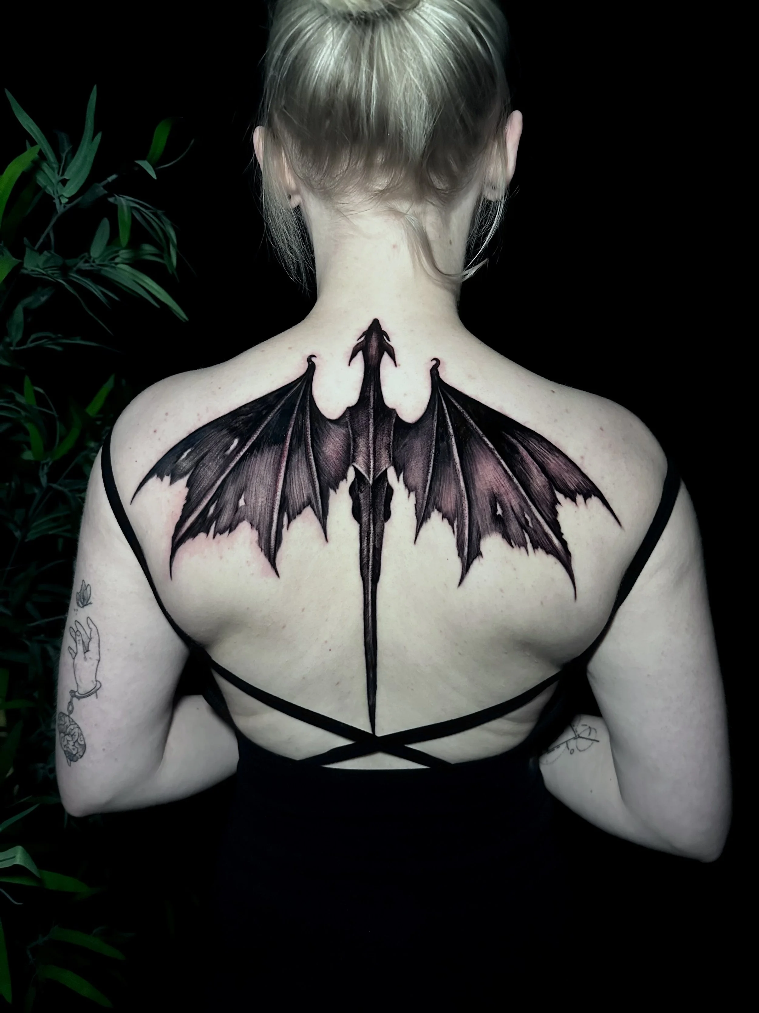 Back view of a person with blonde hair, showcasing a large butterfly tattoo on their upper back and shoulders, with dark wings and detailed patterns, set against a black background with some green plant leaves visible on the left side.