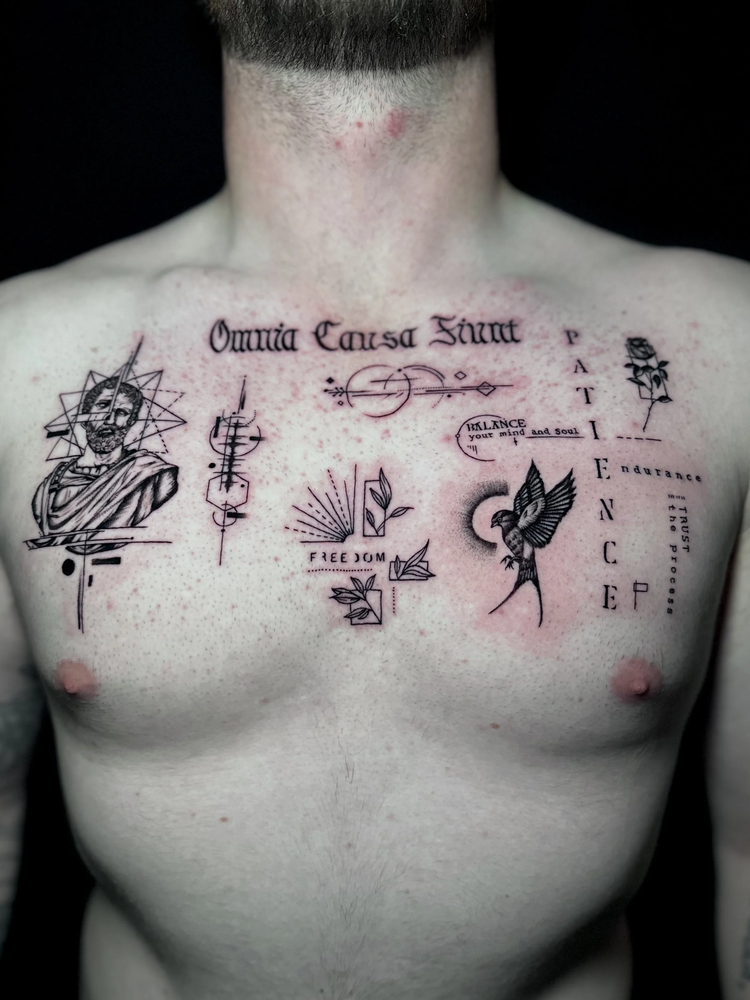 Close-up of a man's chest with various tattoo designs and words, including a portrait of Jesus, geometric patterns, and text in Latin.