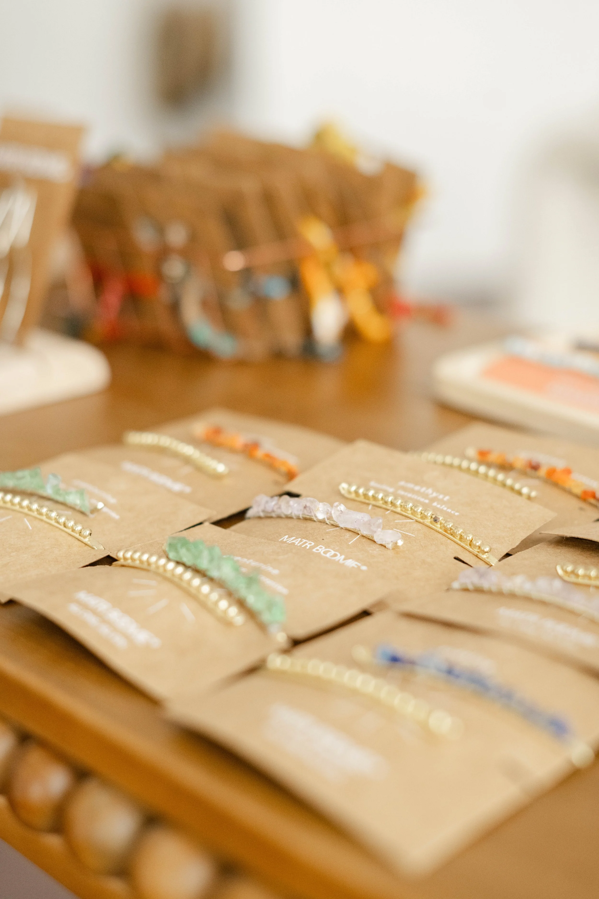 Display of handmade jewelry on cardboard cards, arranged on a wooden surface. The jewelry includes beaded necklaces and bracelets in various colors.