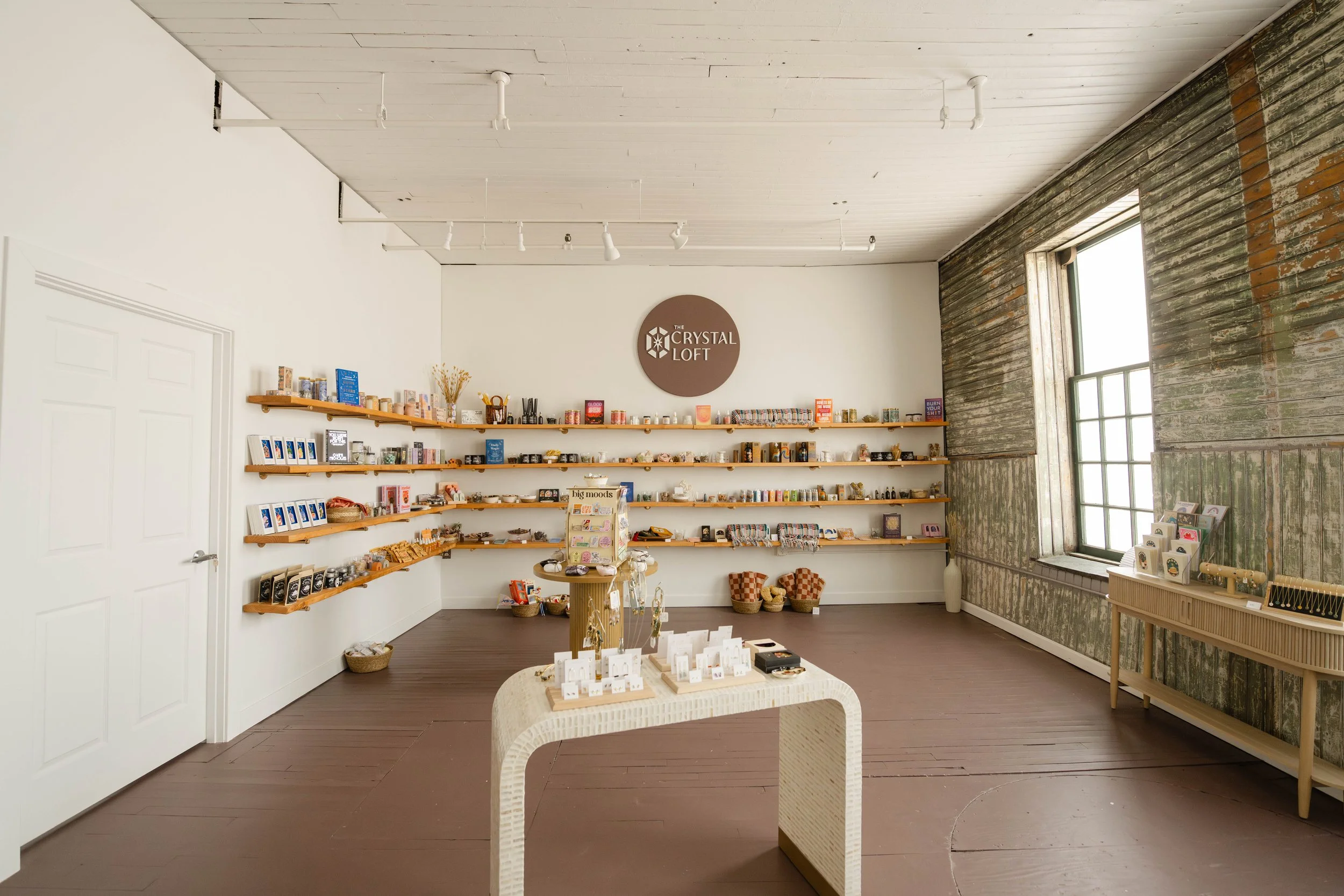 Interior of a retail store called The Crystal Loft with wooden shelves displaying jewelry, books, and decorative items, large windows, and a white ceiling with track lighting.