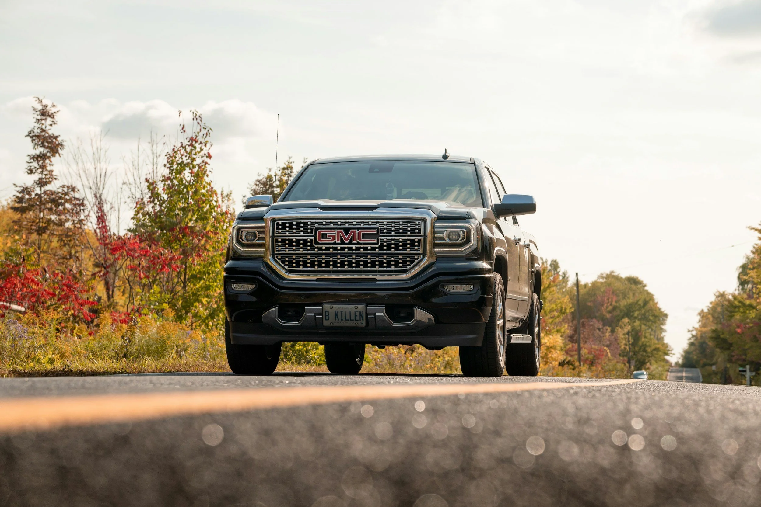 A black GMC pickup truck parked on a road, with colorful autumn trees and a cloudy sky in the background.