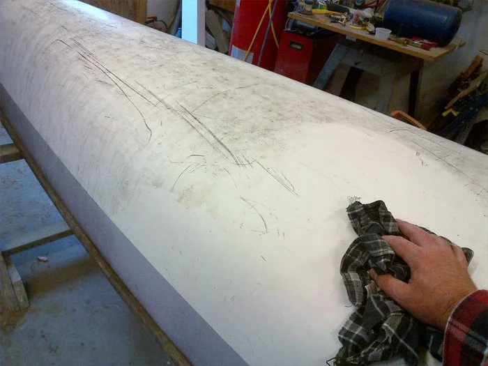 Person's hand cleaning the surface of a large, elongated, white surfboard in a workshop.