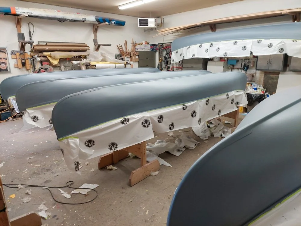 Four kayak hulls painted in light gray color resting on wooden stands in a workshop.