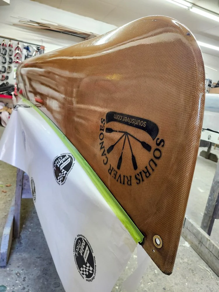 A carbon fiber surfboard with the logo of Souris River Canoe Outfitters and Soustisriver.com, resting on a surface covered with protective paper in a workshop.