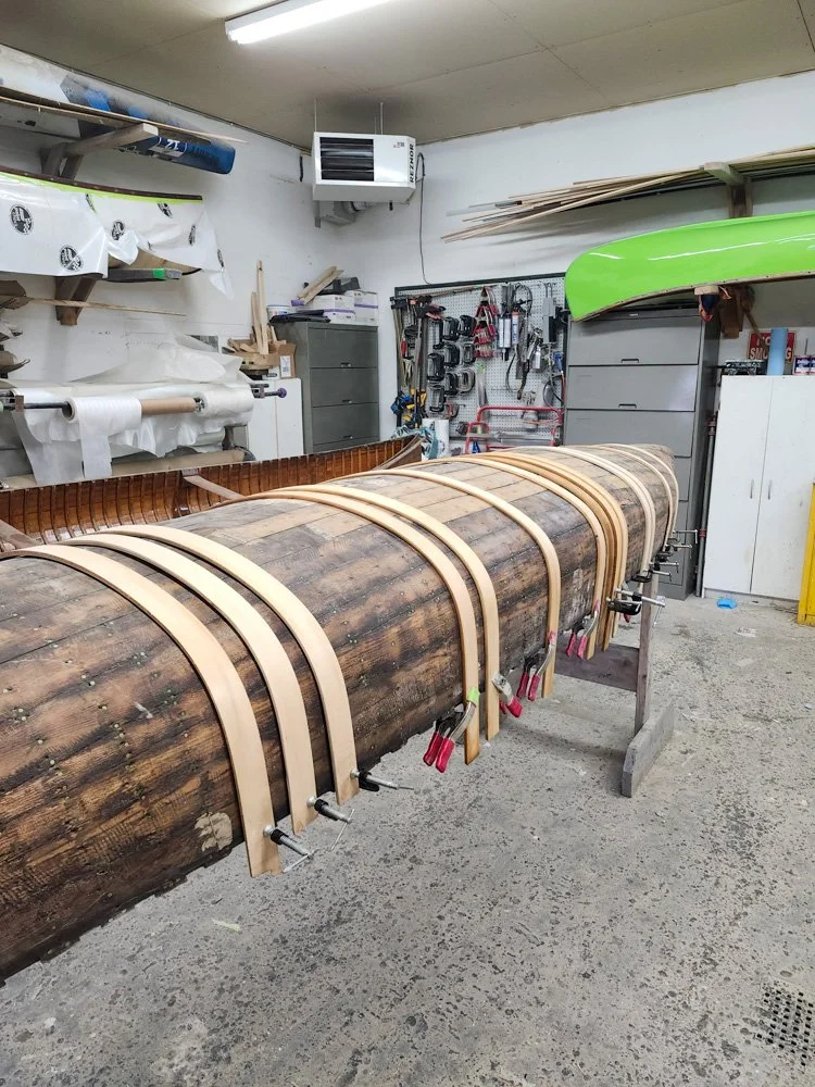 Wooden kayak or canoe in a workshop, secured with clamps, with tools and materials in the background.