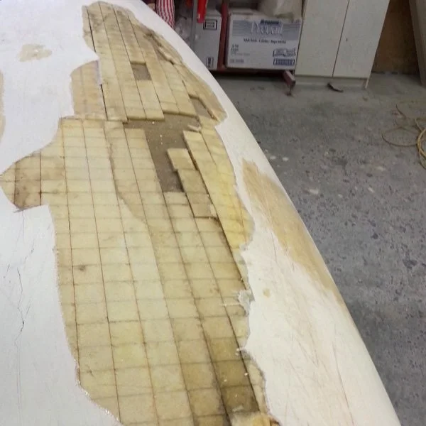 A damaged white boat hull with broken fiberglass and a grid pattern underneath, in a workshop with boxes and tools in the background.
