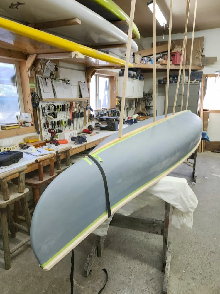 A workshop with a surfboard in the foreground, which is partially painted and secured with straps. Several surfboards are stored on shelves above, and the workspace has tools, equipment, and pegboards on the walls.