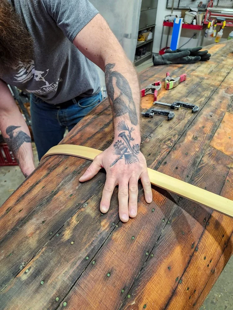 A person with tattoos on their arm and hand working on a wooden workbench. The person is holding a light-colored wooden strip, possibly a bow, in a workshop with tools and supplies in the background.