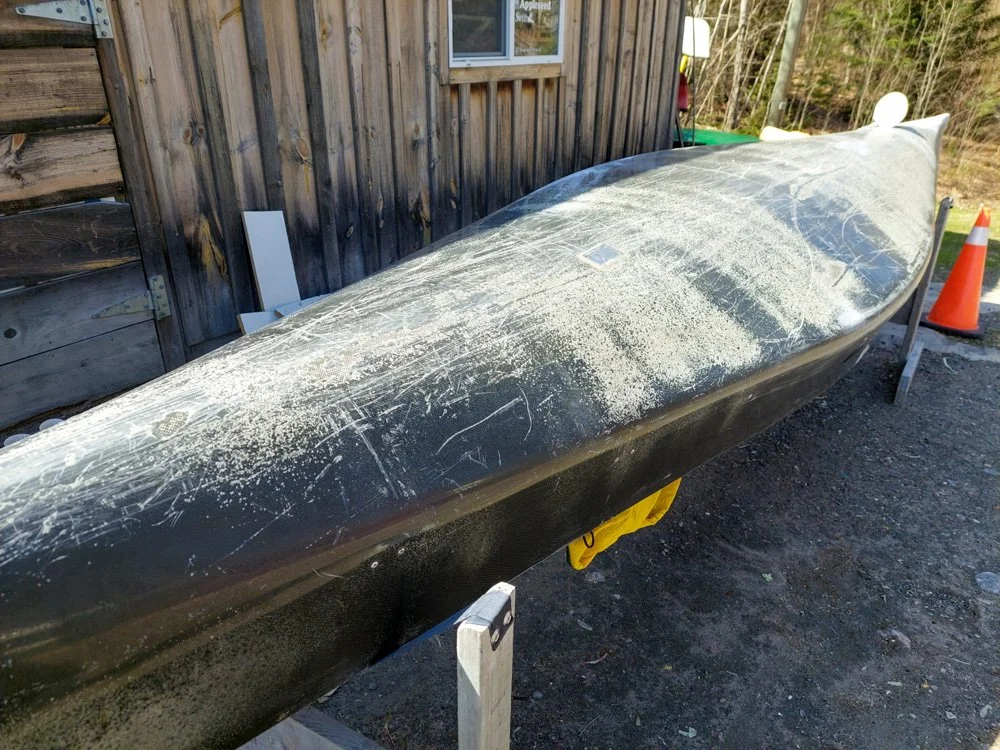 A black kayak with scratches and scuffs on its surface, resting on a wooden stand outside near a wooden shed with a window. An orange traffic cone is visible in the background.