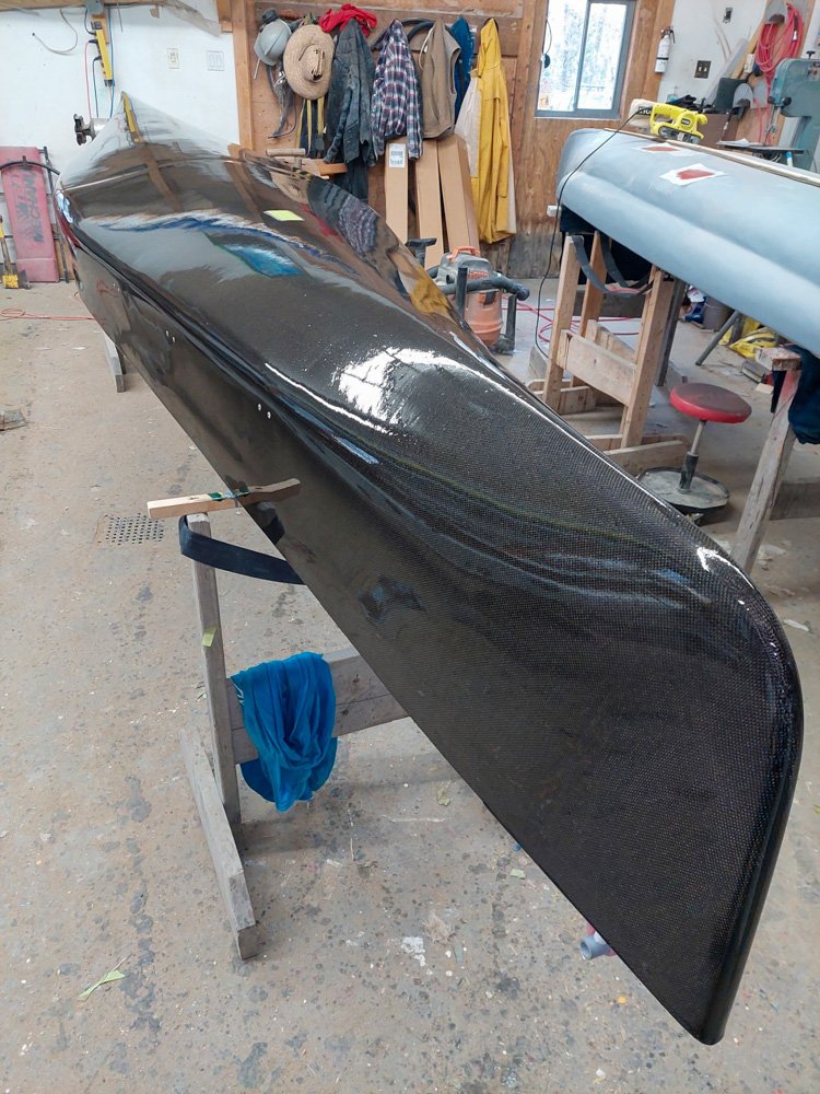 A carbon fiber boat hull on a workbench in a workshop, with tools and clothing hanging in the background.
