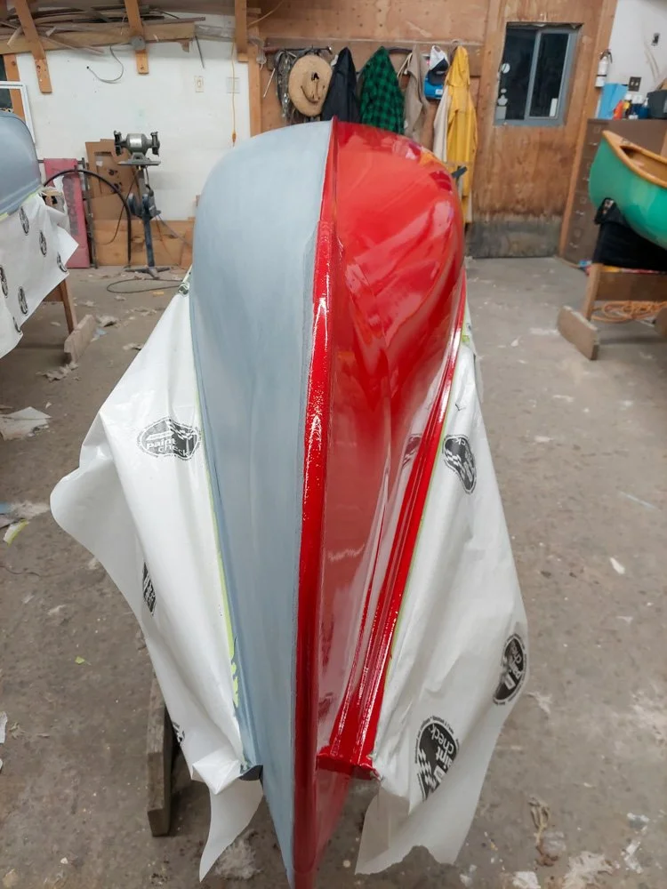A boat in the process of being painted, with the bow covered in a shiny red color and the rest primed with gray primer, situated in a workshop with tools and jackets hanging on the wall.