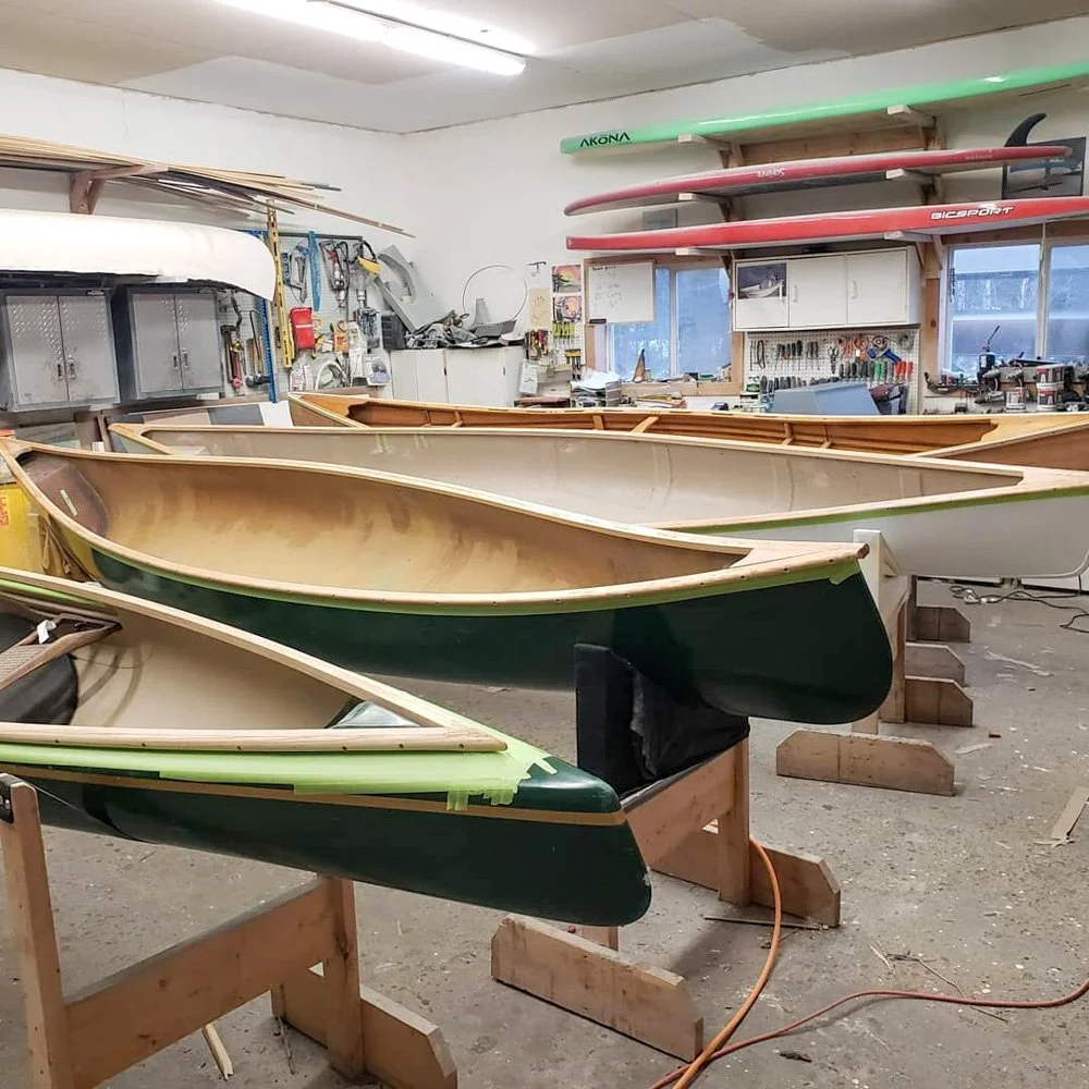 Several canoes in different stages of construction inside a workshop, with tools and equipment on the walls and shelves, and two red and one green paddle stored on racks.