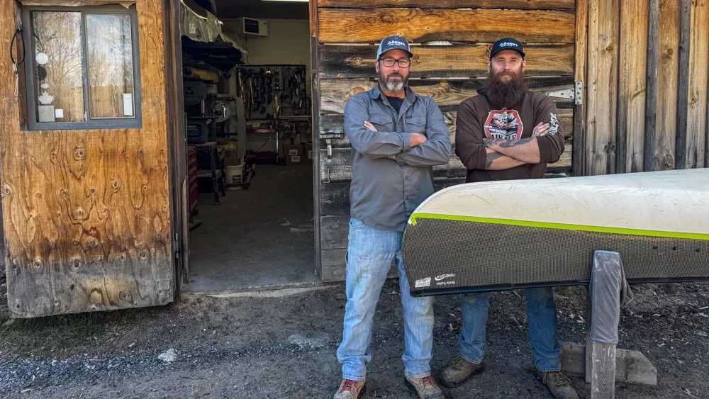 Two men with beards and glasses standing outside a wooden shed, with their arms crossed, posing next to a canoe.