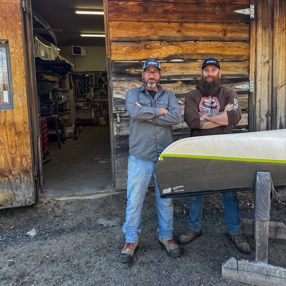 Two men standing outside a wooden workshop with a surfboard in front of them. The man on the left has glasses and a beard, wearing a gray jacket and blue jeans. The man on the right has a full beard and is wearing a brown hoodie featuring a graphic design. The workshop door is open, revealing tools and equipment inside.