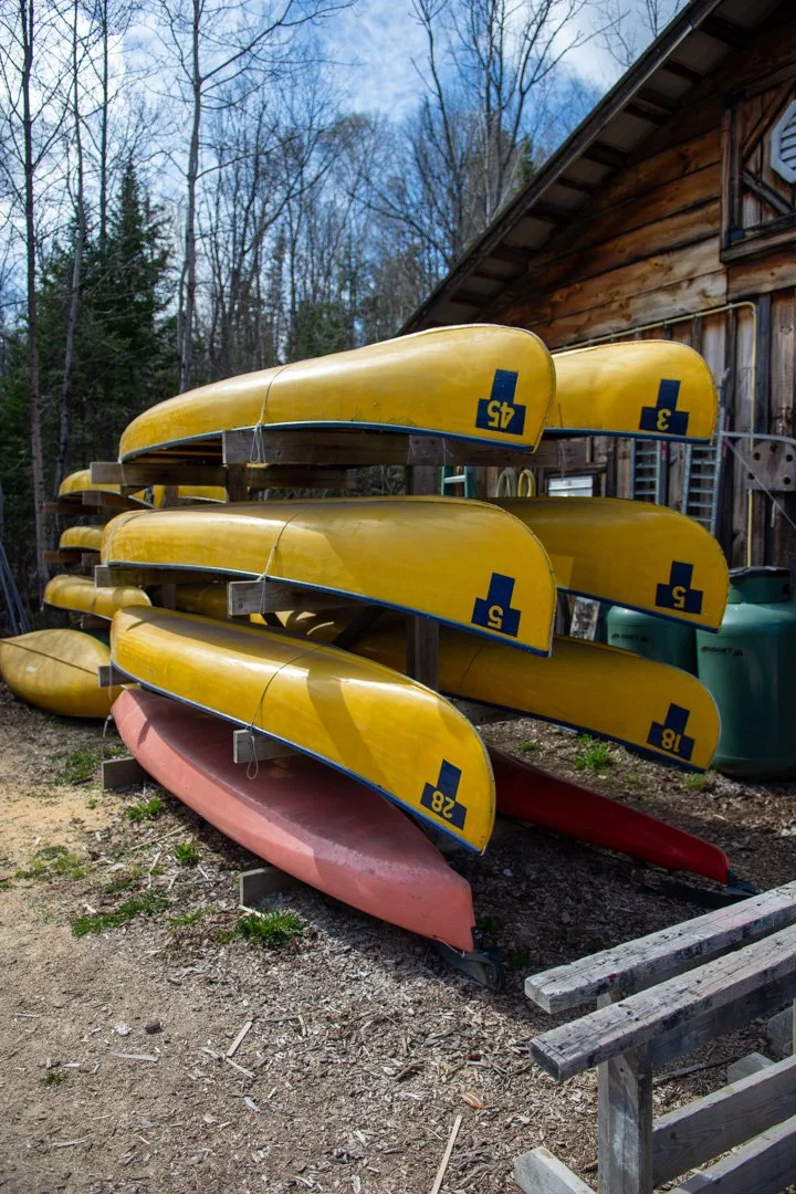 Several yellow canoes and one red canoe stacked on a wooden rack outside near a rustic wooden building, with leafless trees and a partly cloudy sky in the background.