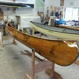 A wooden canoe resting on a stand in a workshop, with another canoe and various tools in the background.