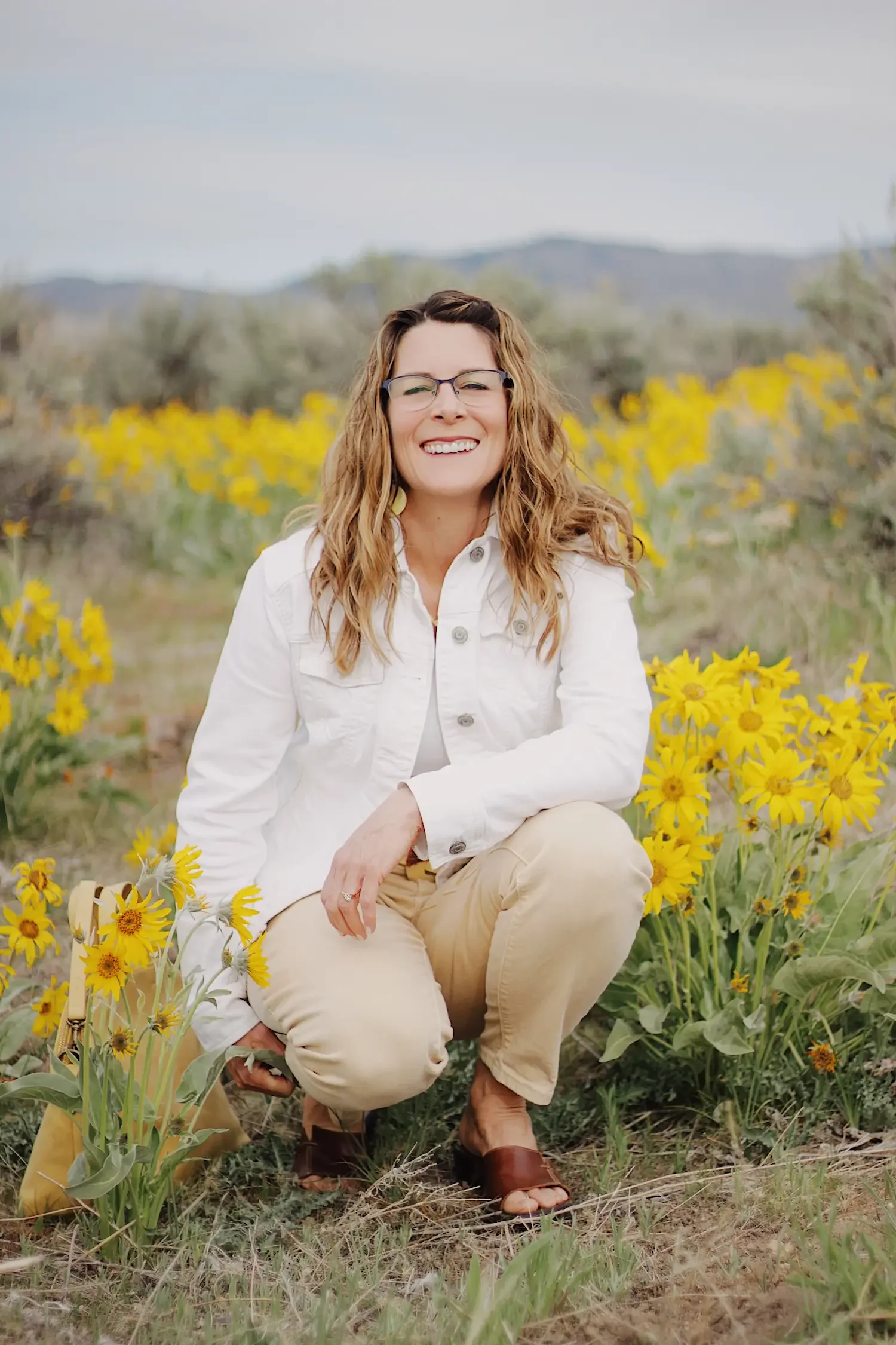 Michelle Batten, LPC, Hopeful Connections Counseling, Boise, Idaho,  with curly hair, glasses, and a white jacket sitting among yellow flowers in a field, smiling at the camera with a mountain backdrop  provides couples counseling.