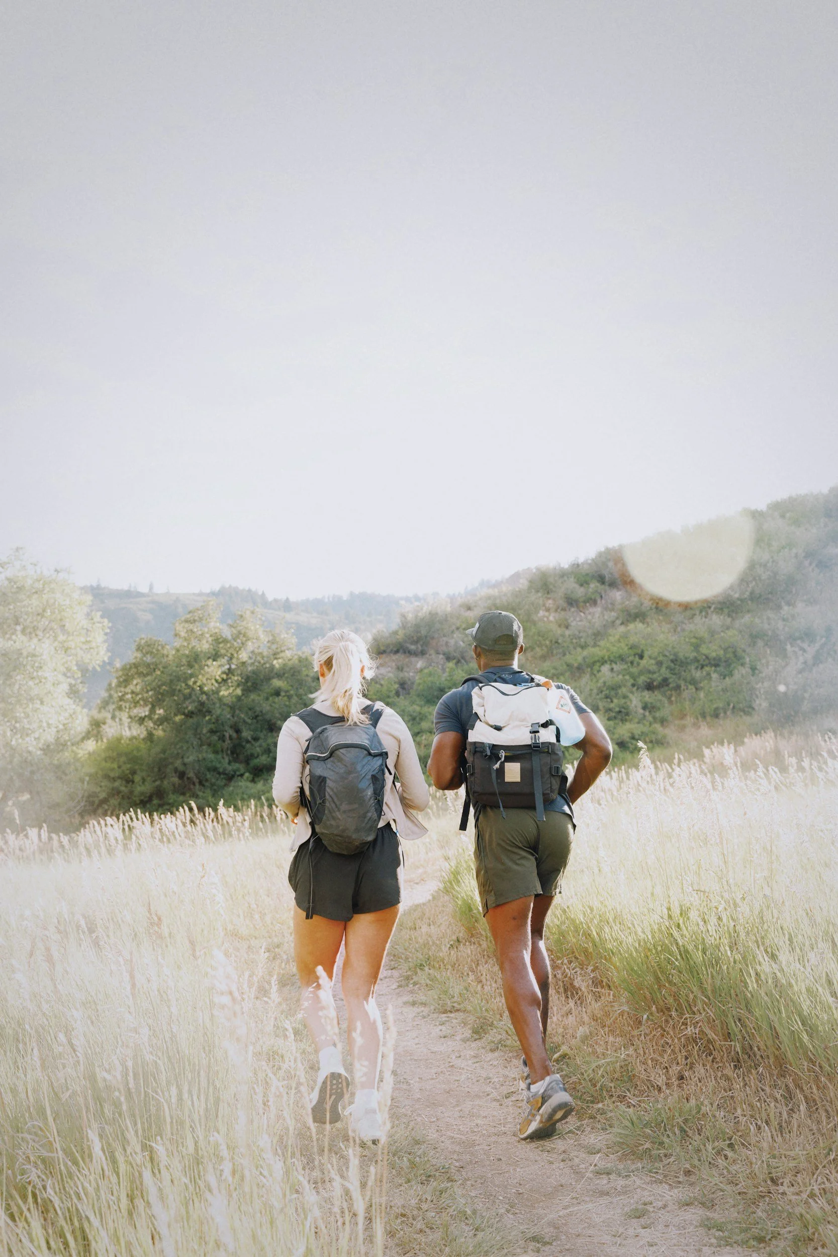 A man and a woman hiking on a trail through a grassy landscape with hills and trees in the background, both wearing backpacks.