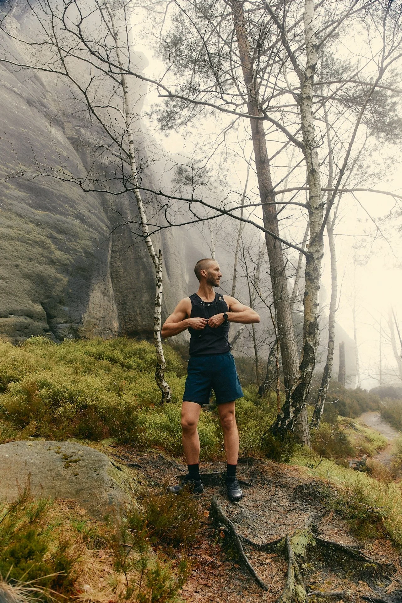 A man standing outdoors in a forested area with bare trees and mossy rocks, wearing a black sleeveless shirt, shorts, and trail running shoes, adjusting his shirt as he looks to the side.