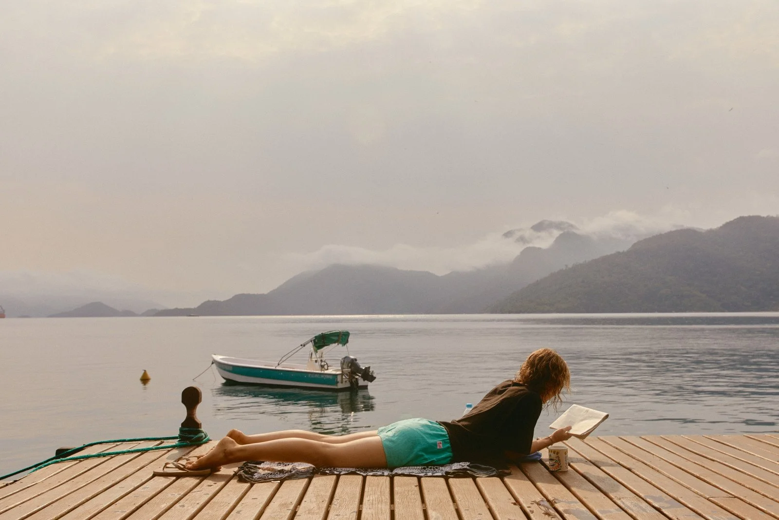 A person with curly hair is lying on a wooden dock, reading a book, beside a serene lake with a small motorboat floating nearby and mountains in the background.