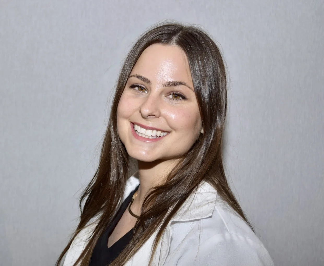 A young woman with long brown hair smiling, wearing a white top and black shirt, standing against a plain light gray background.