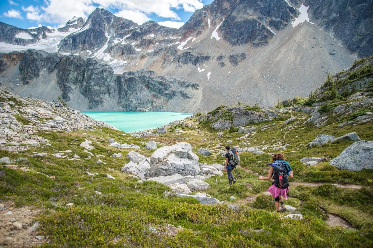 Two hikers walking along a trail in a mountainous area with snow-capped peaks, a turquoise lake, and green vegetation.