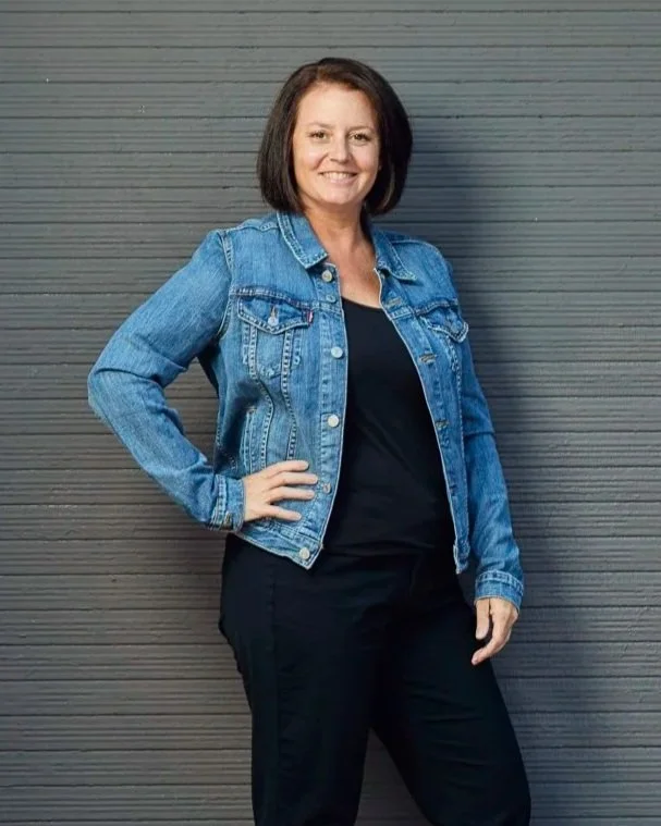 Erin Rue, a confident CPA for small businesses, stands relaxed against a textured gray wall, wearing a denim jacket and black outfit, ready to inspire tax confidence.