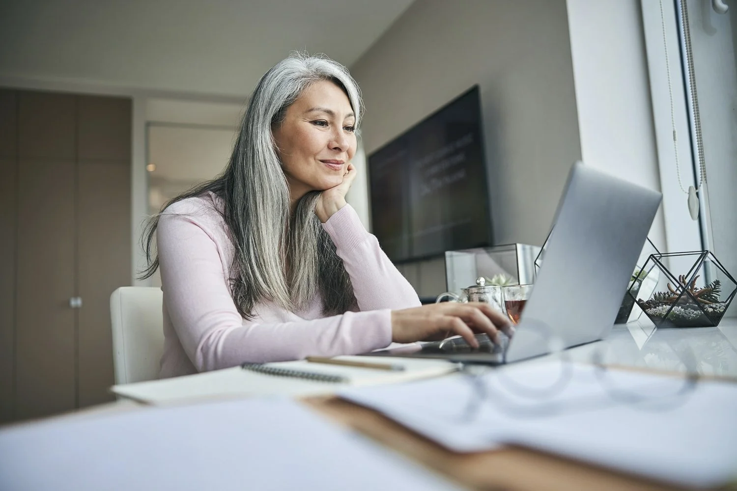 A woman with long gray hair sitting at a desk, using a laptop, in a bright office space.