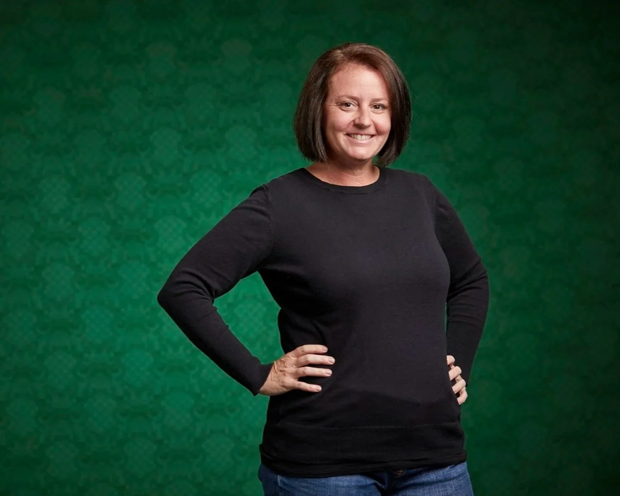 Erin Rue exudes confidence in a black sweater and jeans, standing against a vibrant green backdrop, ready to inspire small business owners as their trusted tax accountant.