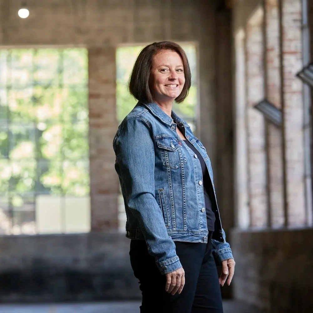 Erin Rue, tax accountant for small businesses, a white woman with shoulder-length brown hair, wearing a denim jacket and black pants, smiling in an industrial-style space with large windows and brick walls.