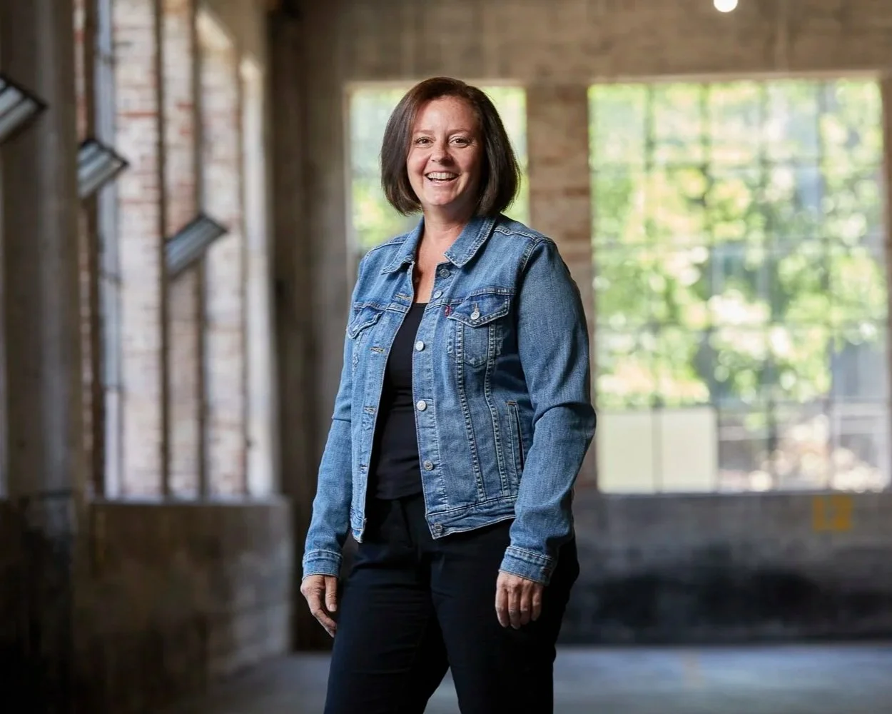 Erin Rue, tax accountant for small businesses, a white woman with shoulder-length brown hair, wearing a denim jacket and black pants, smiling in an industrial-style space with large windows and brick walls.