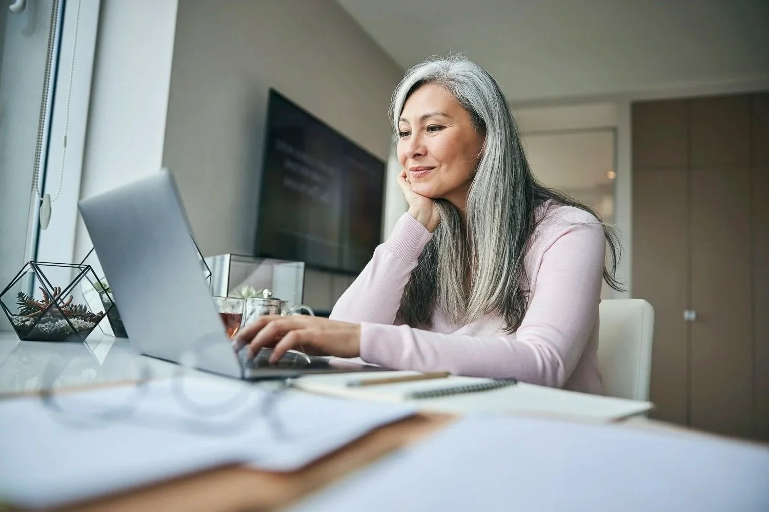 A happy, silver-haired woman in a cozy pink sweater smiles while working on her laptop, surrounded by notebooks and a glass of tea, embodying the confidence and ease of small business tax preparation.