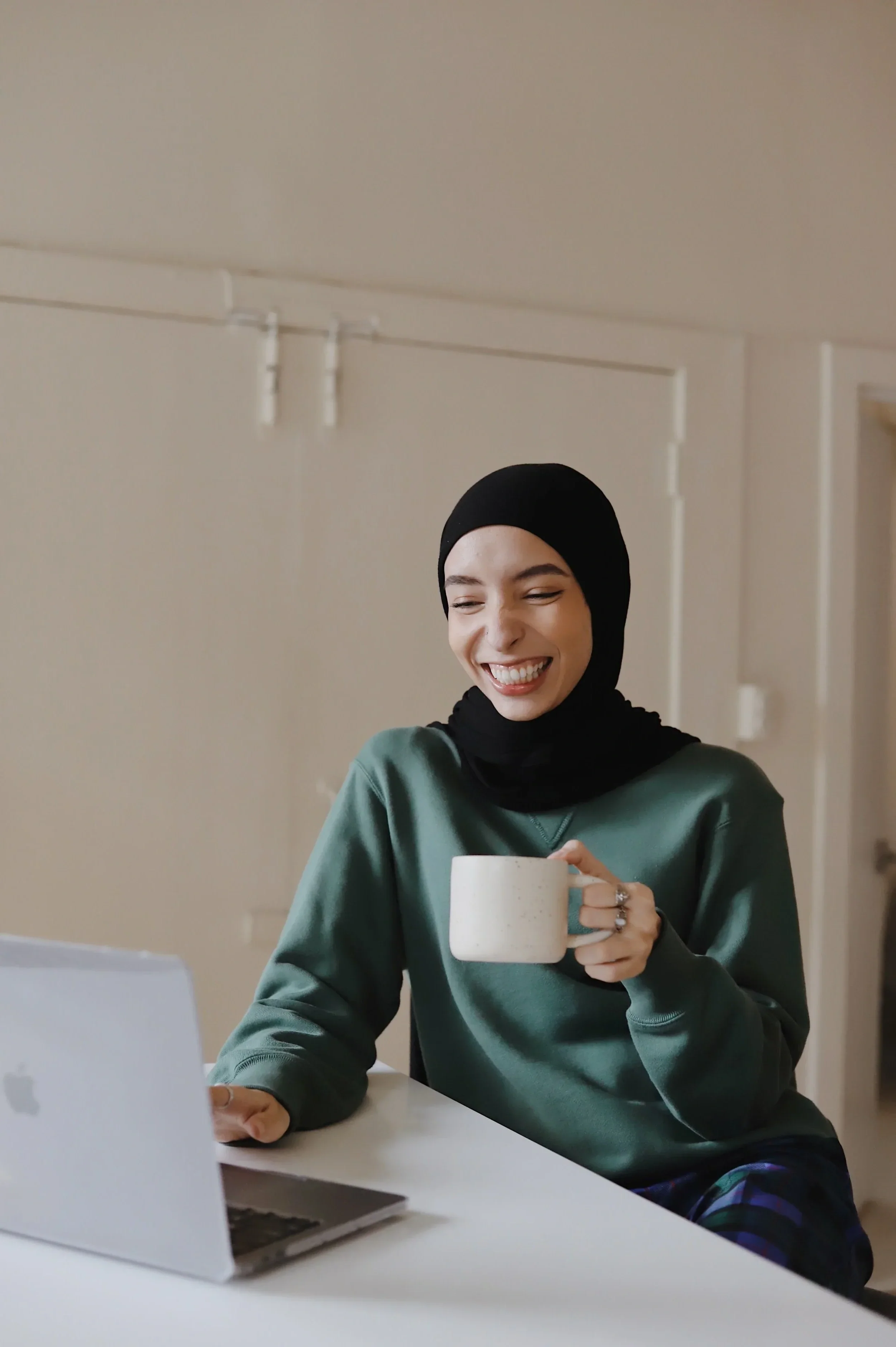 Smiling woman in a hijab sitting at a desk in front of a laptop, holding a mug, in a bright modern room.